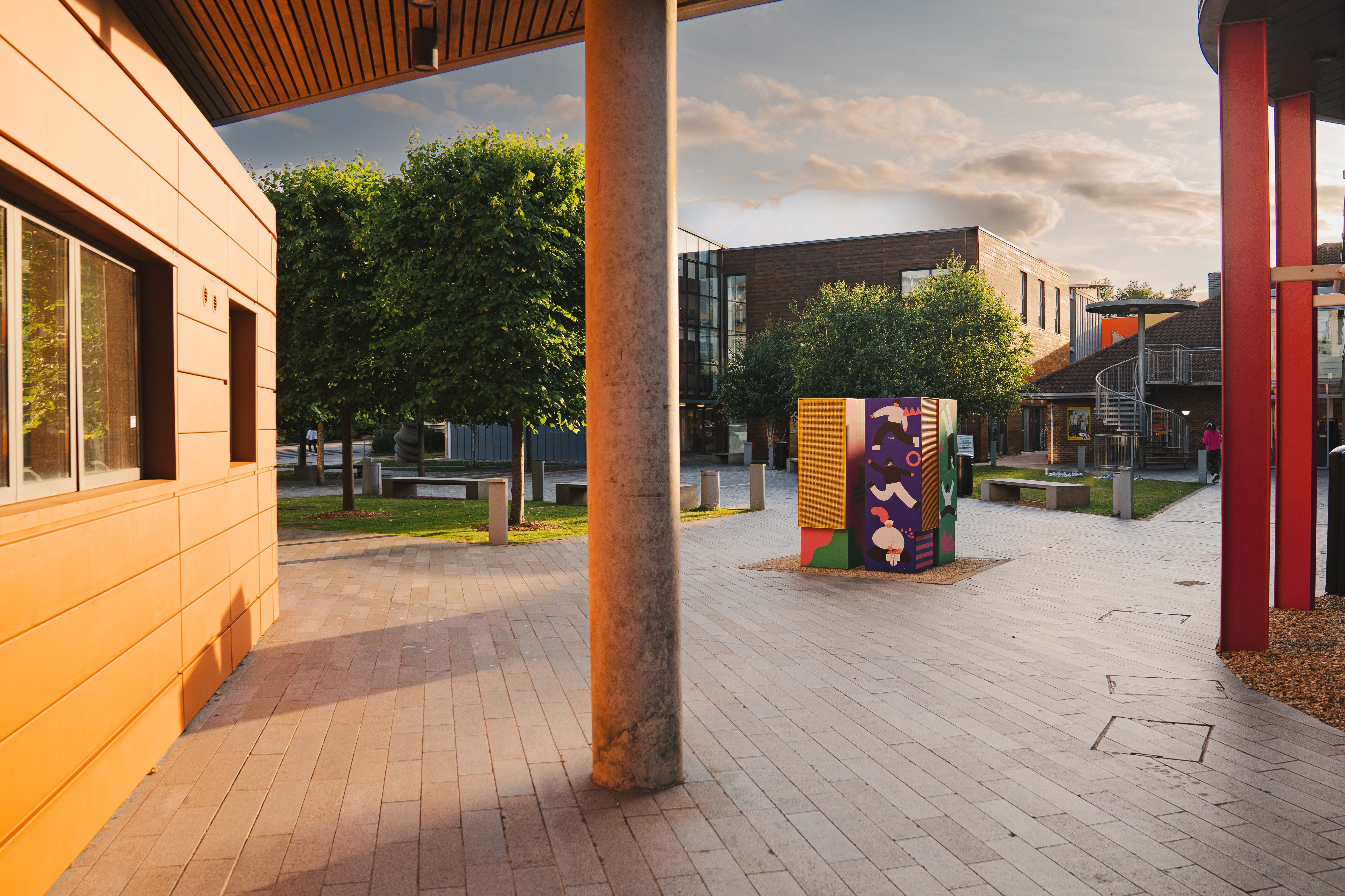 Modern courtyard with timber-clad building, red metal columns, paved plaza, purple artwork installation, and contemporary architecture.