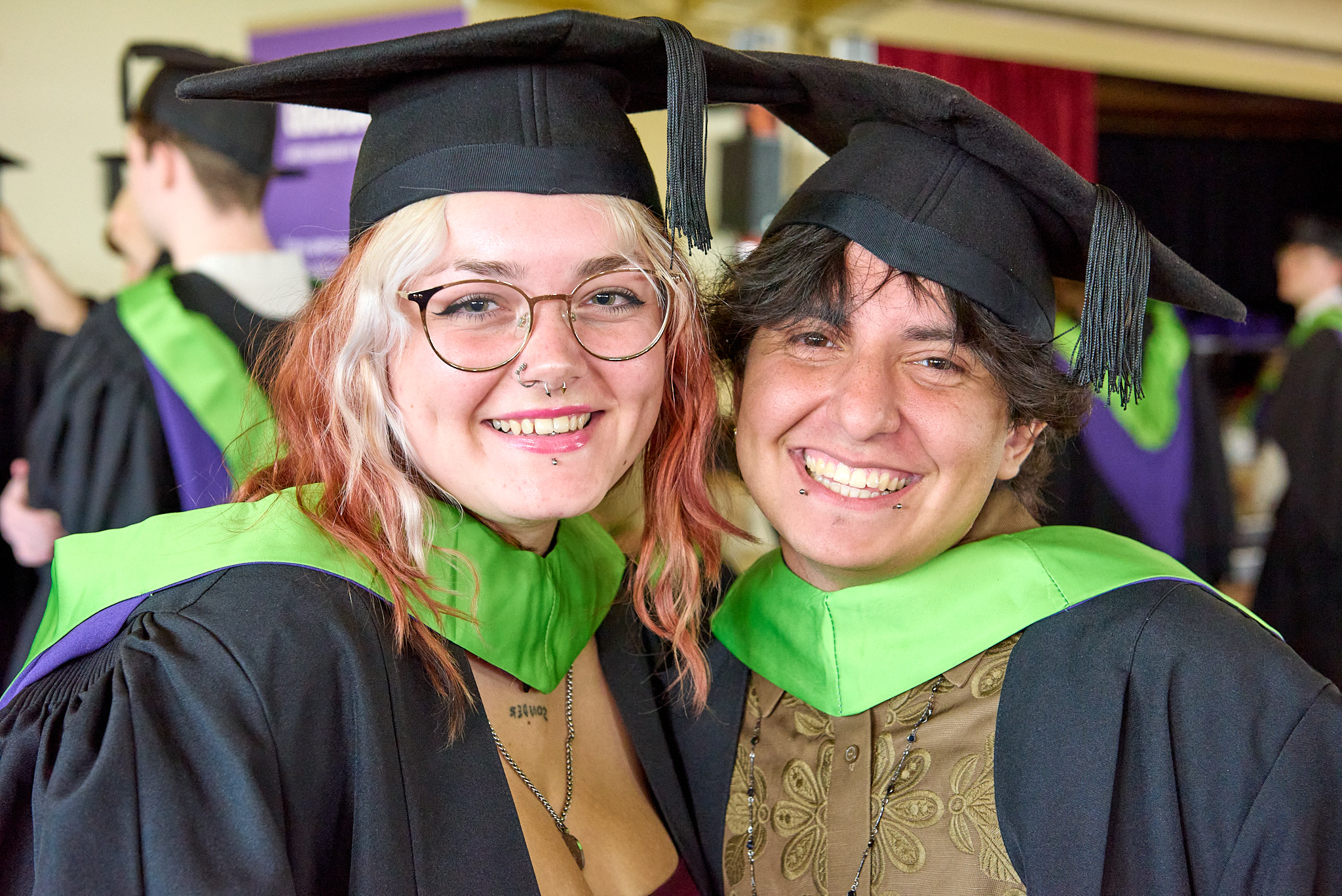 Two graduates in black caps and gowns with bright green stoles stand together indoors, with other graduates blurred in the background.