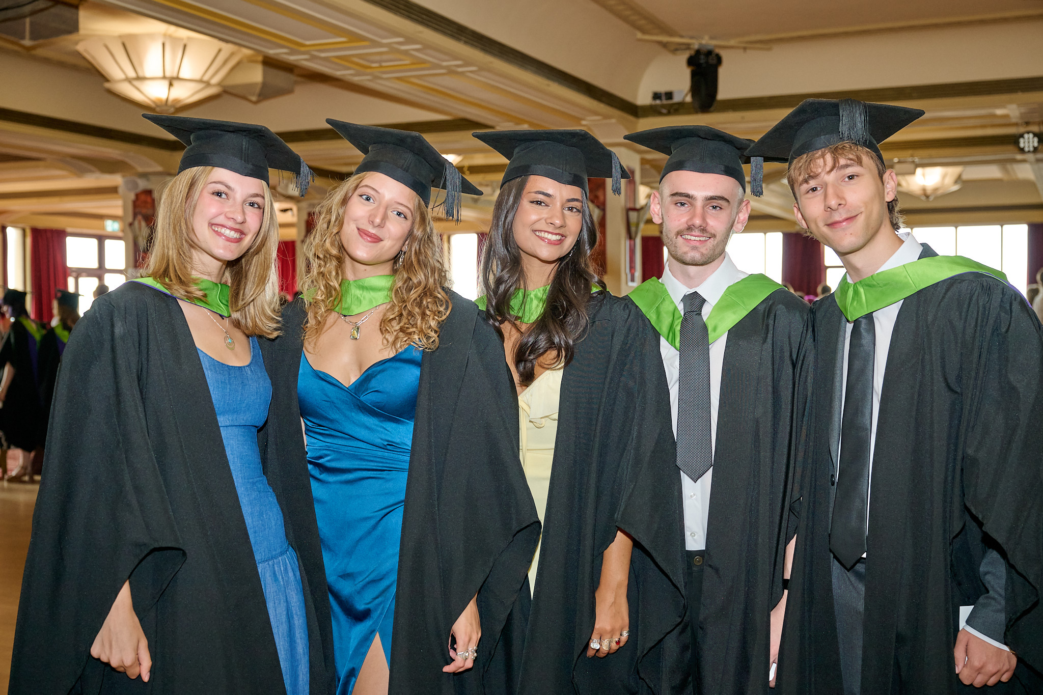 Five graduates in caps and gowns stand together in a decorated indoor hall with warm lighting.