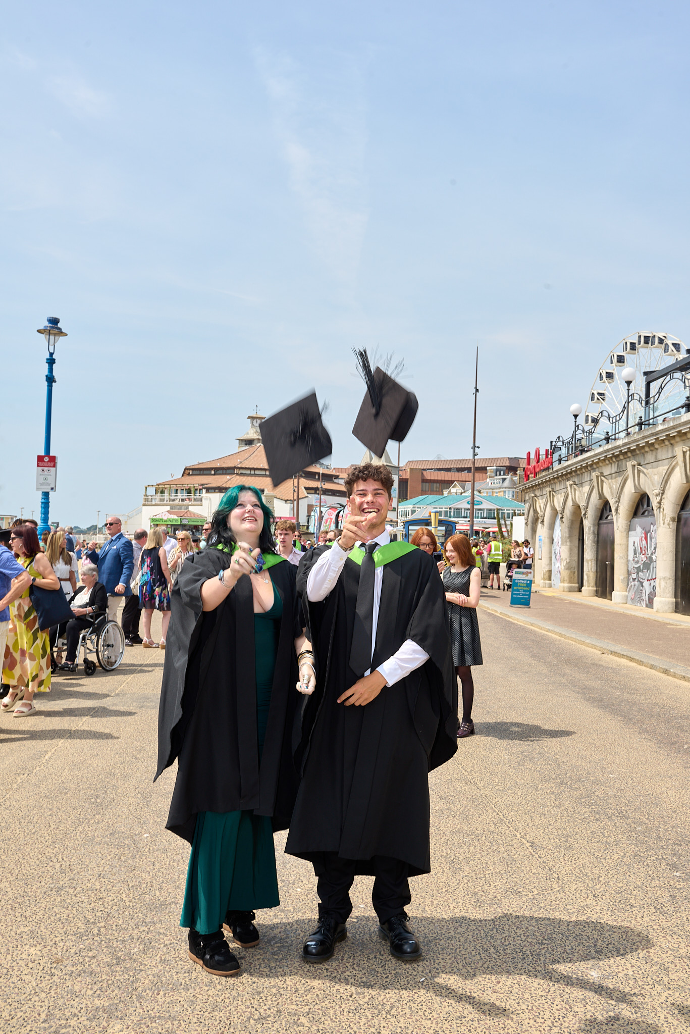 Two graduates in black gowns and mortarboards stand on a seaside promenade, with other people and a pier with arches in the background.