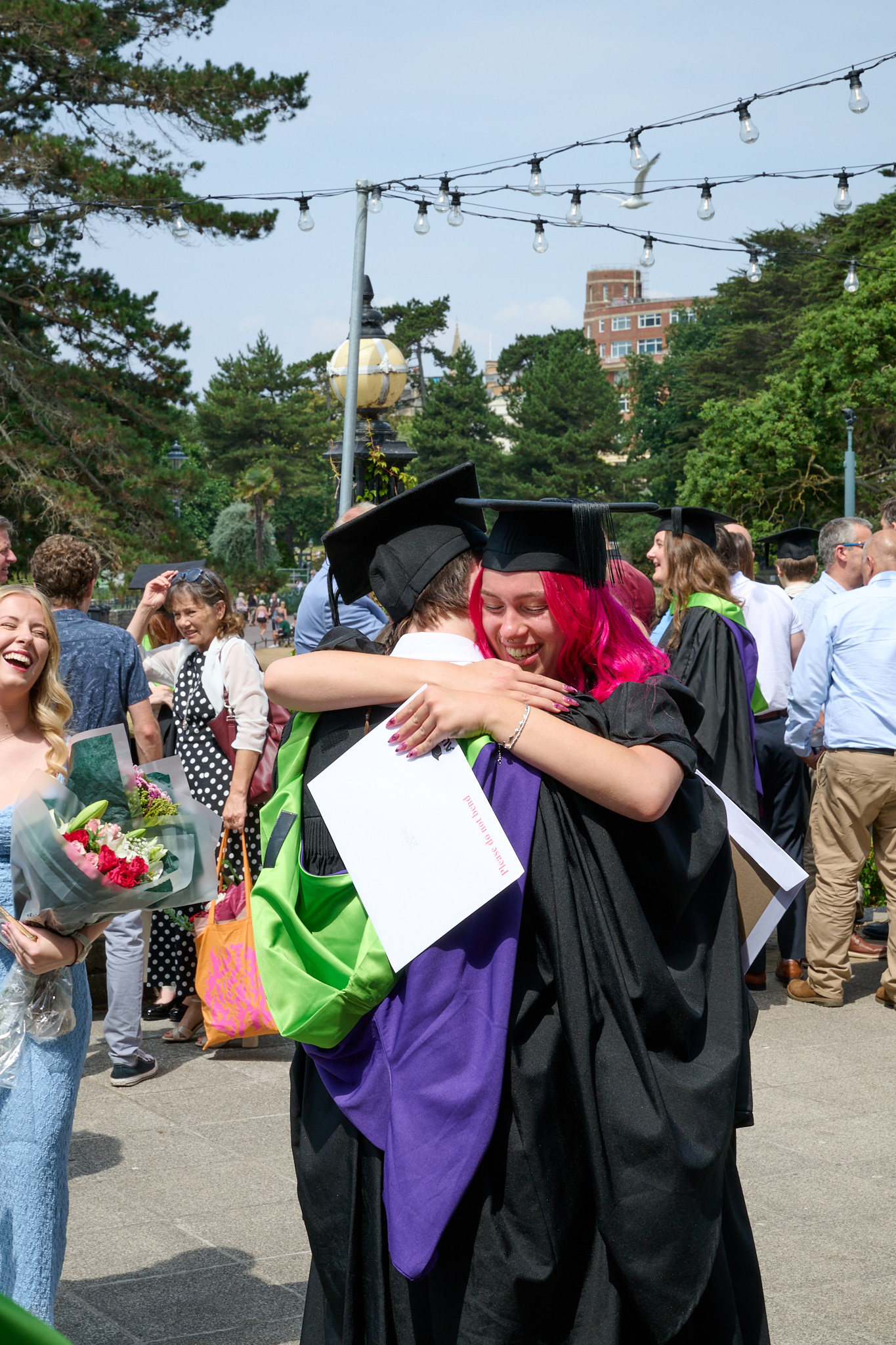 Two graduates in black caps and gowns embrace outdoors, with string lights overhead and people gathered in the background.