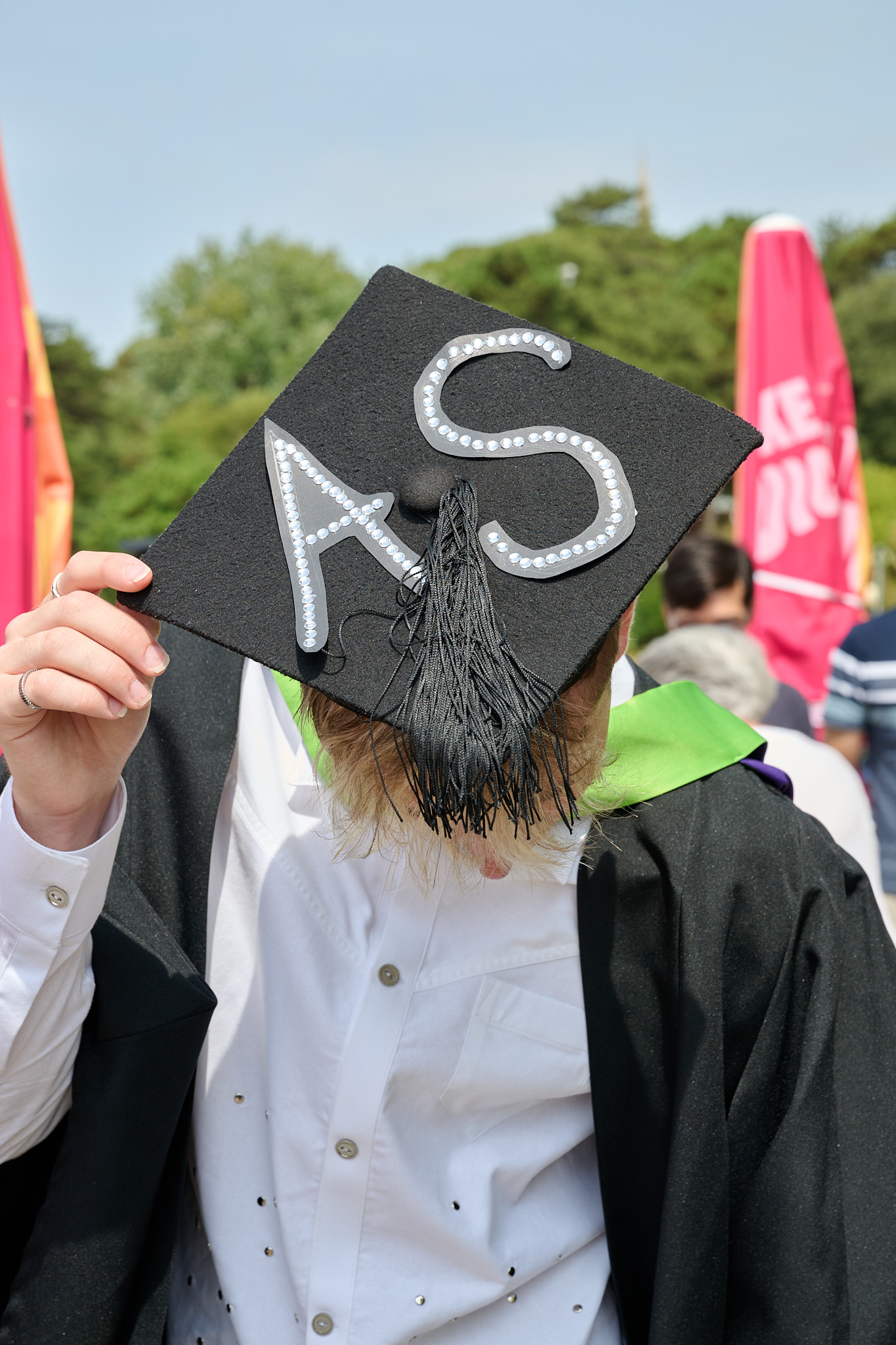 A graduate in a black cap and gown holds the cap over their face, with trees and a pink banner in the background.