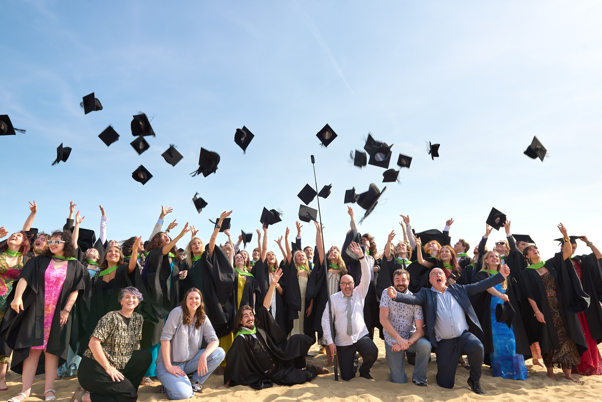 A group of graduates in black gowns and mortarboards pose on a beach as caps fly overhead, with a clear blue sky behind them.