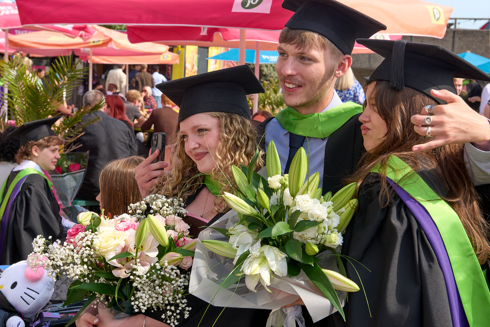 Three graduates in caps and gowns hold bouquets at an outdoor ceremony, with a crowded venue and pink canopy in the background.