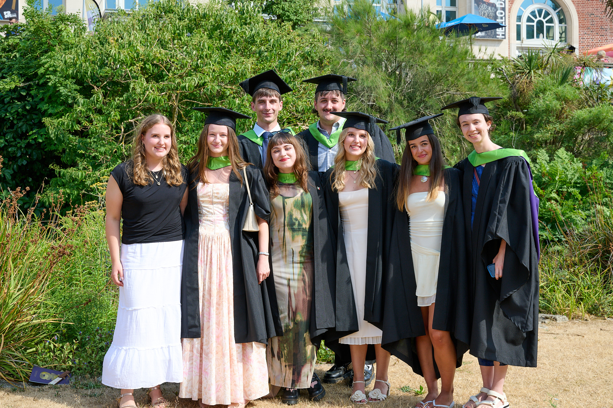 Seven graduates in caps and gowns stand together outdoors in front of greenery and a brick building.