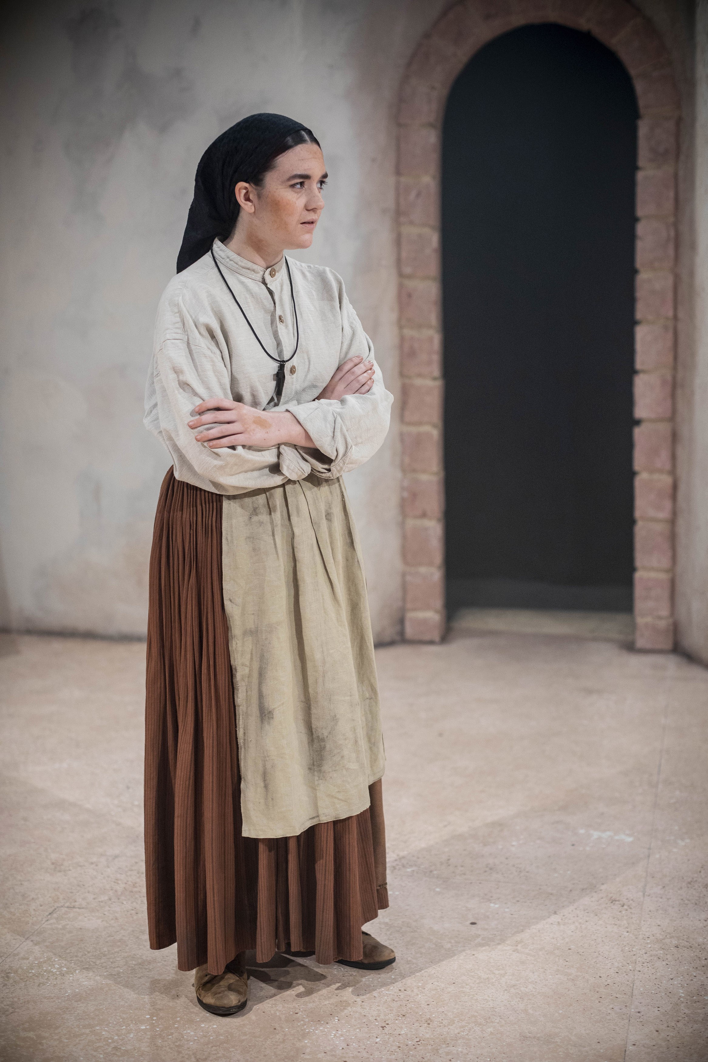 A person in costume stands with their arms folded near a brick arch doorway.