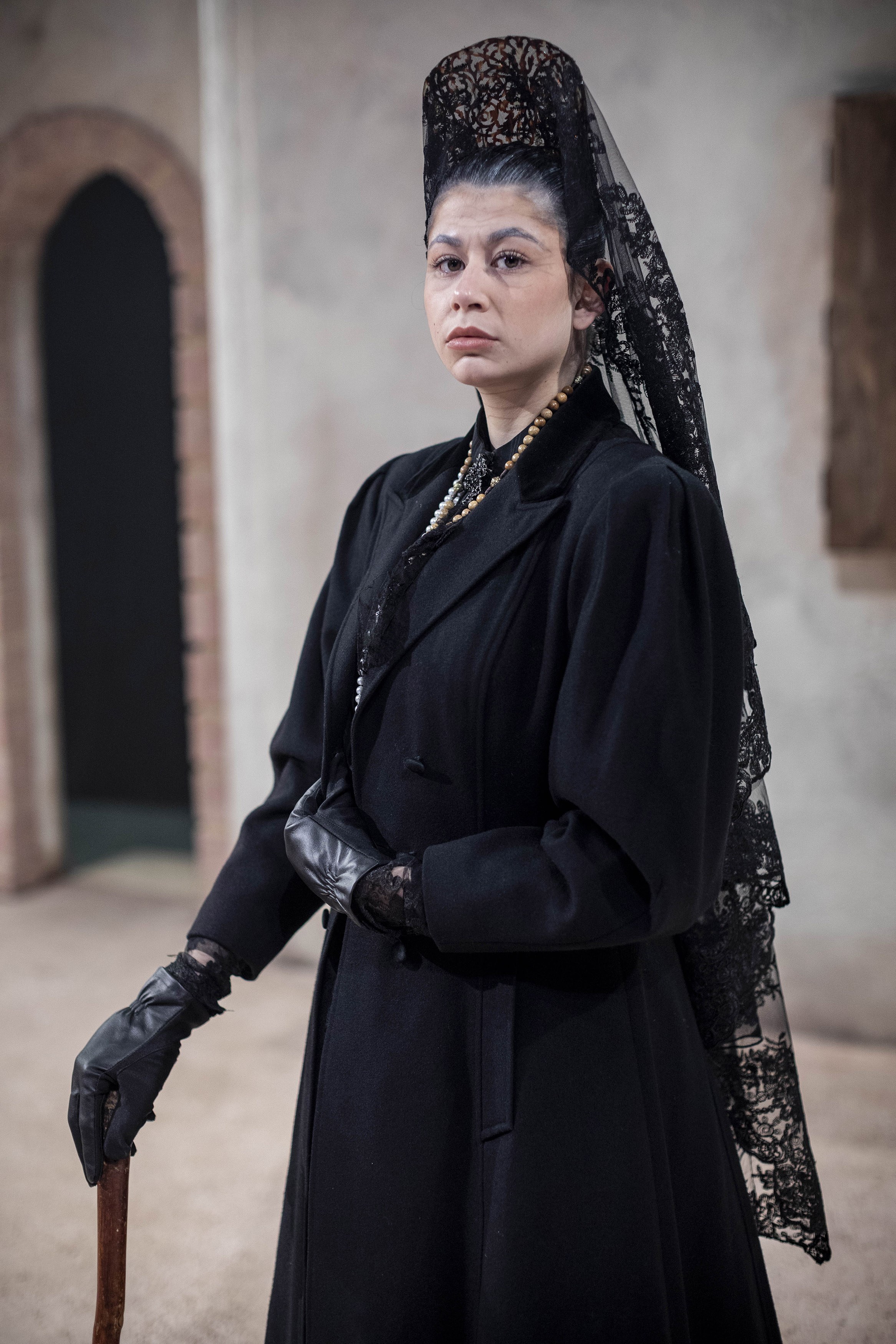 A person in black funeral dress including a long veil and black gloves stands leaning on a cane.