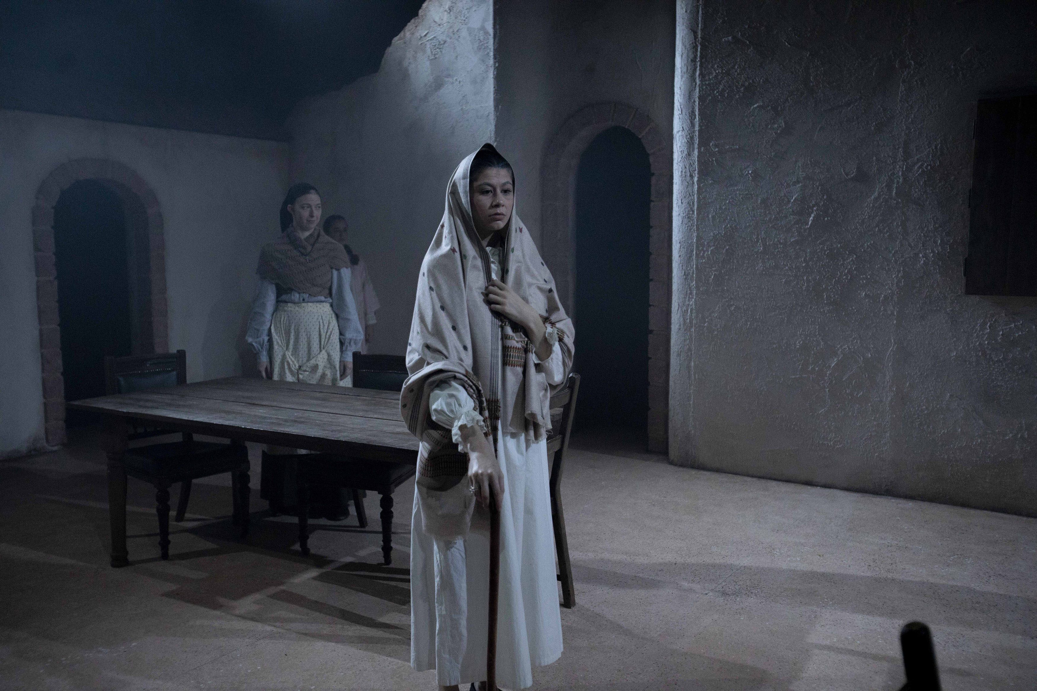 Actors on stage with a dining table. Actor at the front has a shawl draped over their head and is holding a cane.