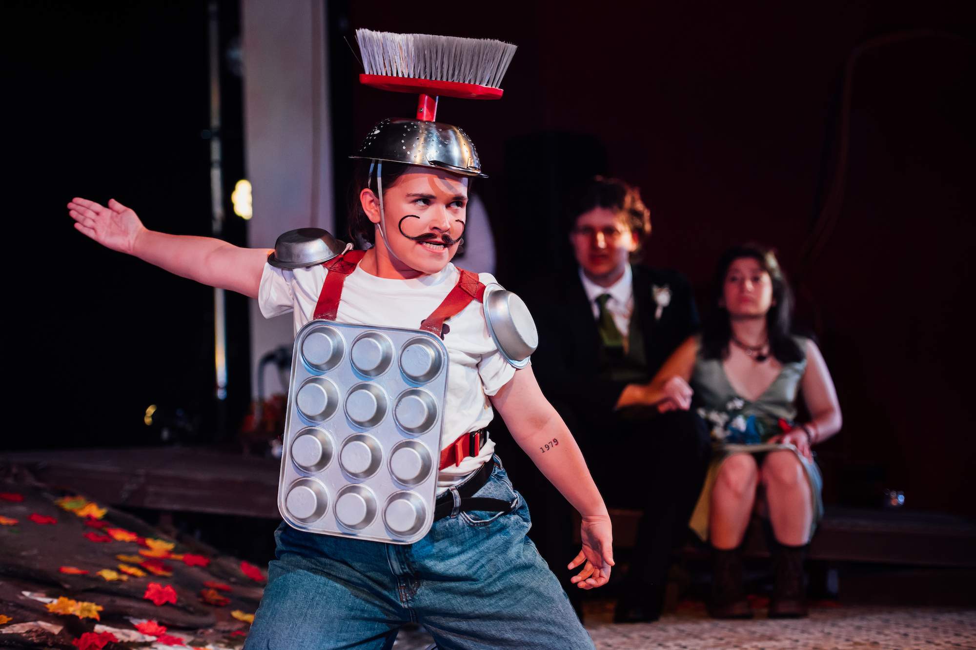 Actor on stage in A Midsummer Night's Dream wearing kitchen utensil costume with red straps and moustache.