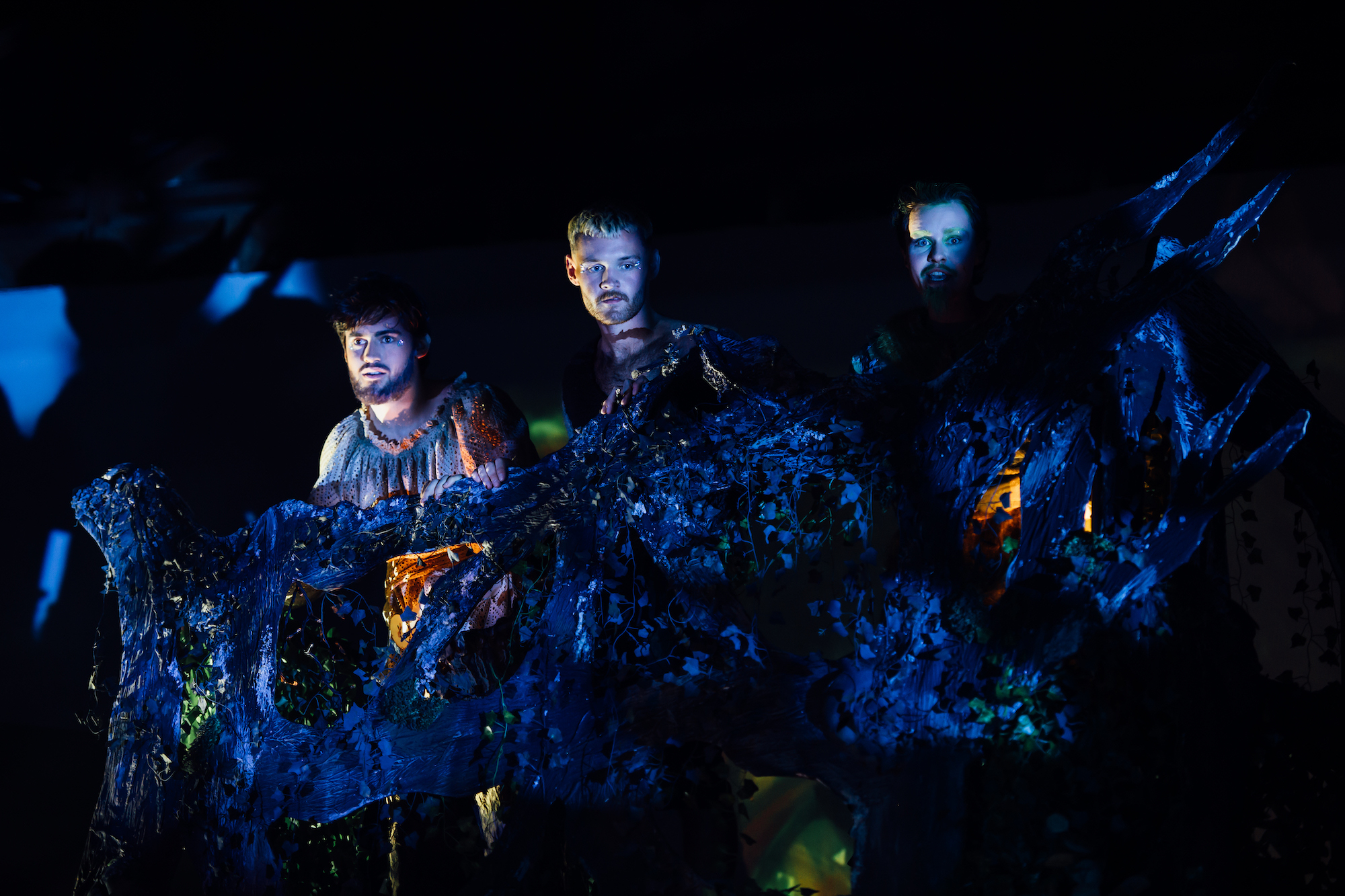 Actors on stage in A Midsummer Night's Dream hiding in a tree, dim blue lighting.