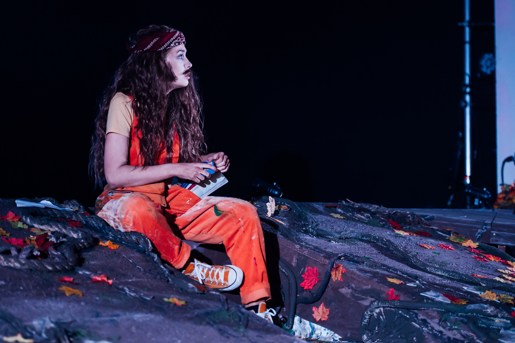 An actor in A Midsummer Night's Dream wearing a red outfit with wavy hippy hair and moustache.