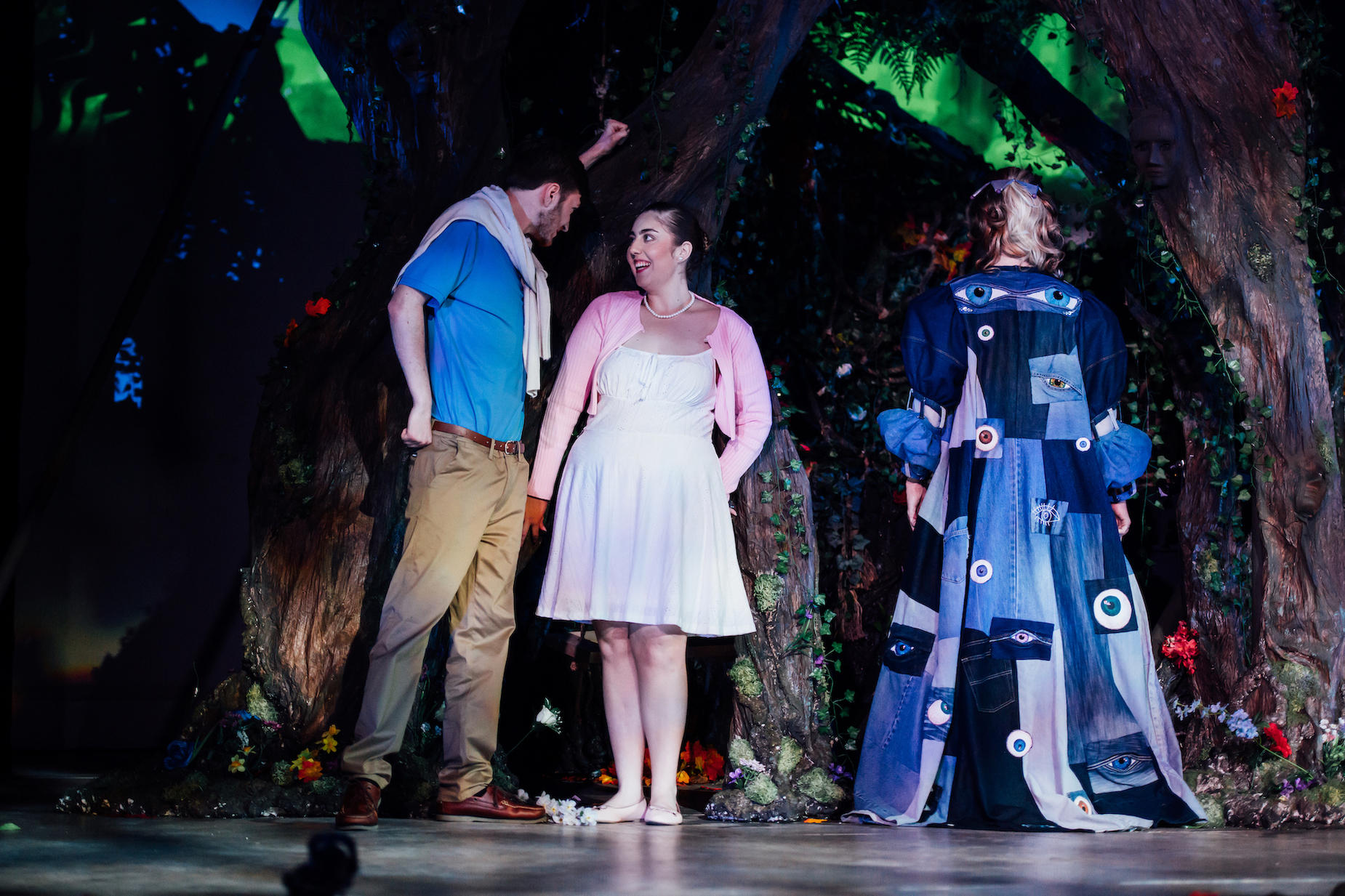 Actors on stage in A Midsummer Night's Dream, two in the woods with trees and a character facing away in a blue eye robe.