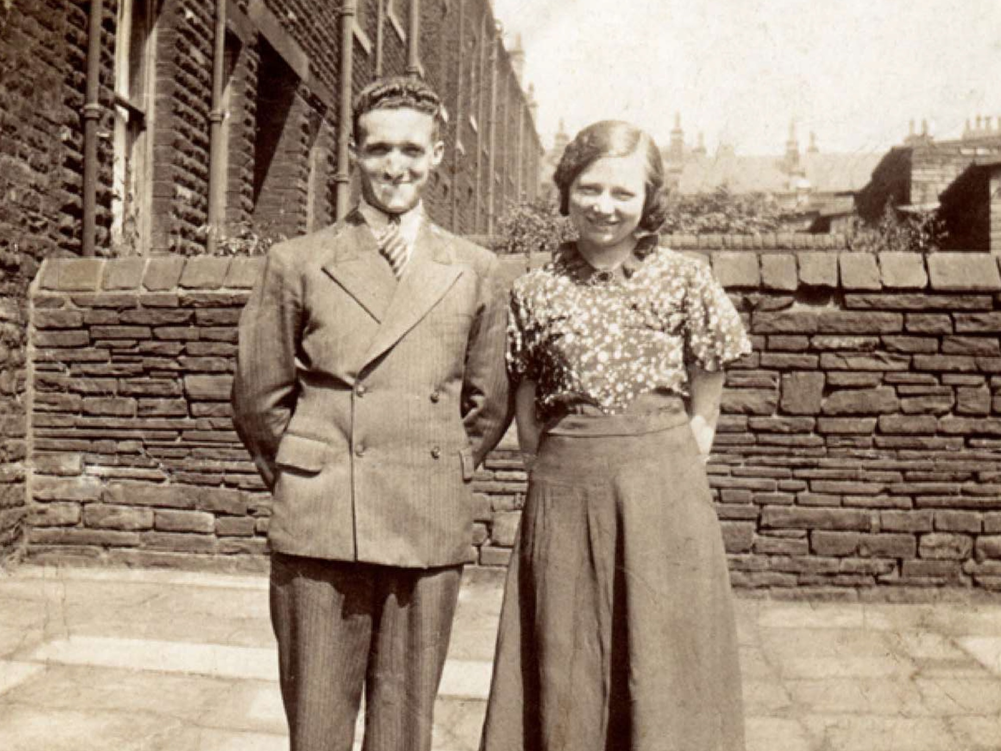 Early 1900s-style sepia photograph of a man and woman smiling with a garden wall behind them.