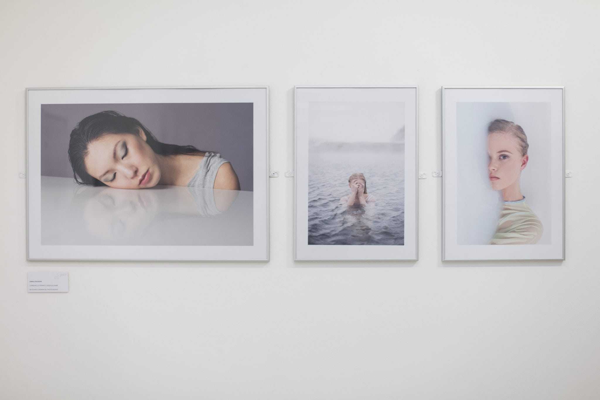 Three photo on a white wall, two images depict two women laying the face on a white object and the third shows a women covering her face as she swims in the ocean