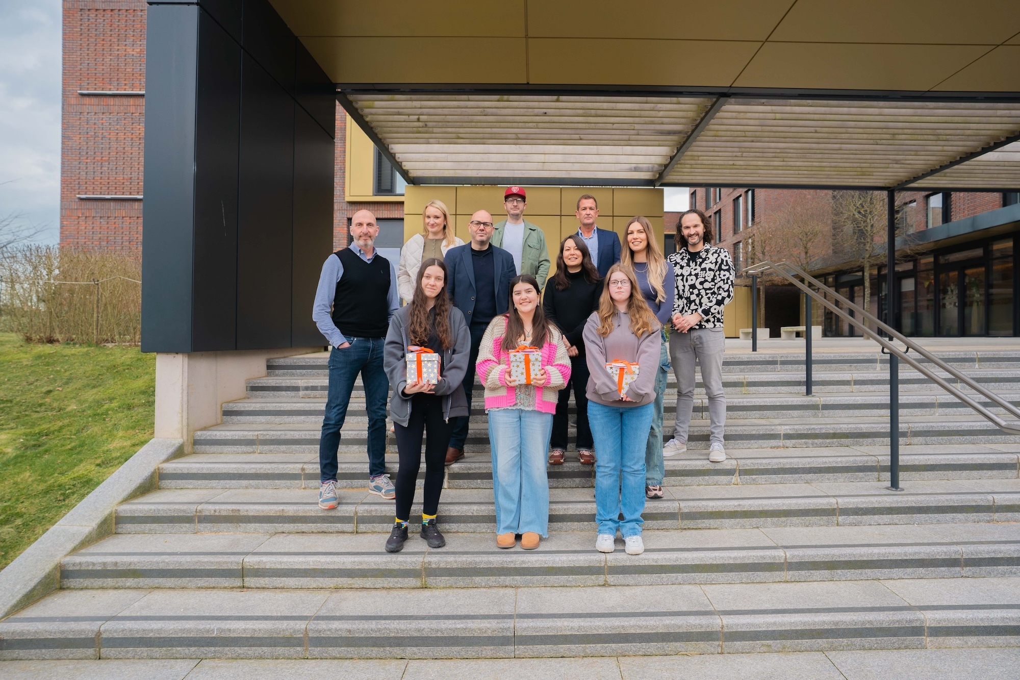 R Melmoth-Bennett, M Smith and A. Tandy posing with wrapped boxes styled like presents, posing for a photo with a team of people from The Creative Collab on the steps outside Campus Halls, Arts University Bournemouth.