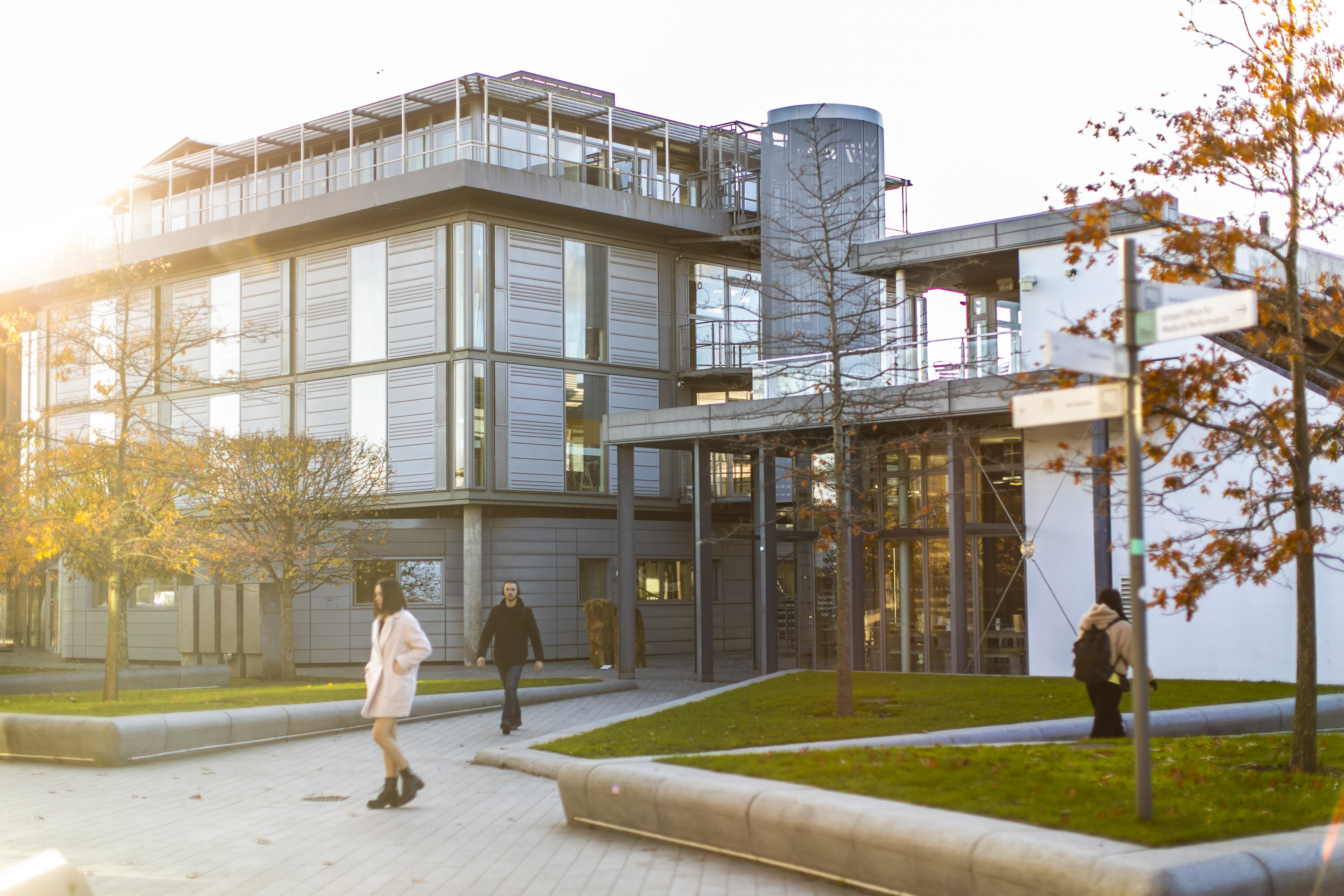 Two buildings with metal and glass exteriors stood in a courtyard with grass verges and trees. People are walking through the courtyard.