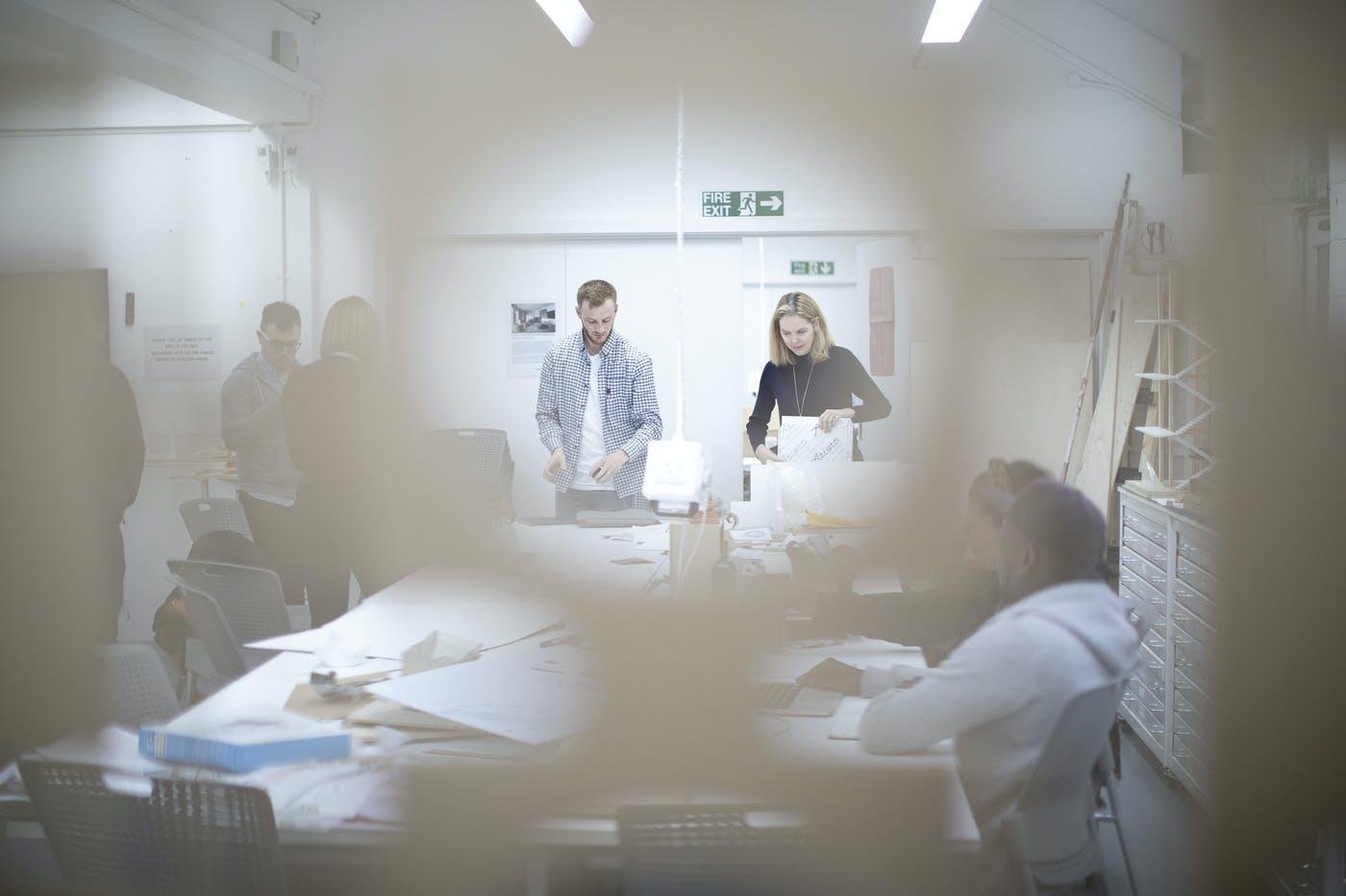 Two people standing at the head of a long table in a studio, other people sat around the table working.