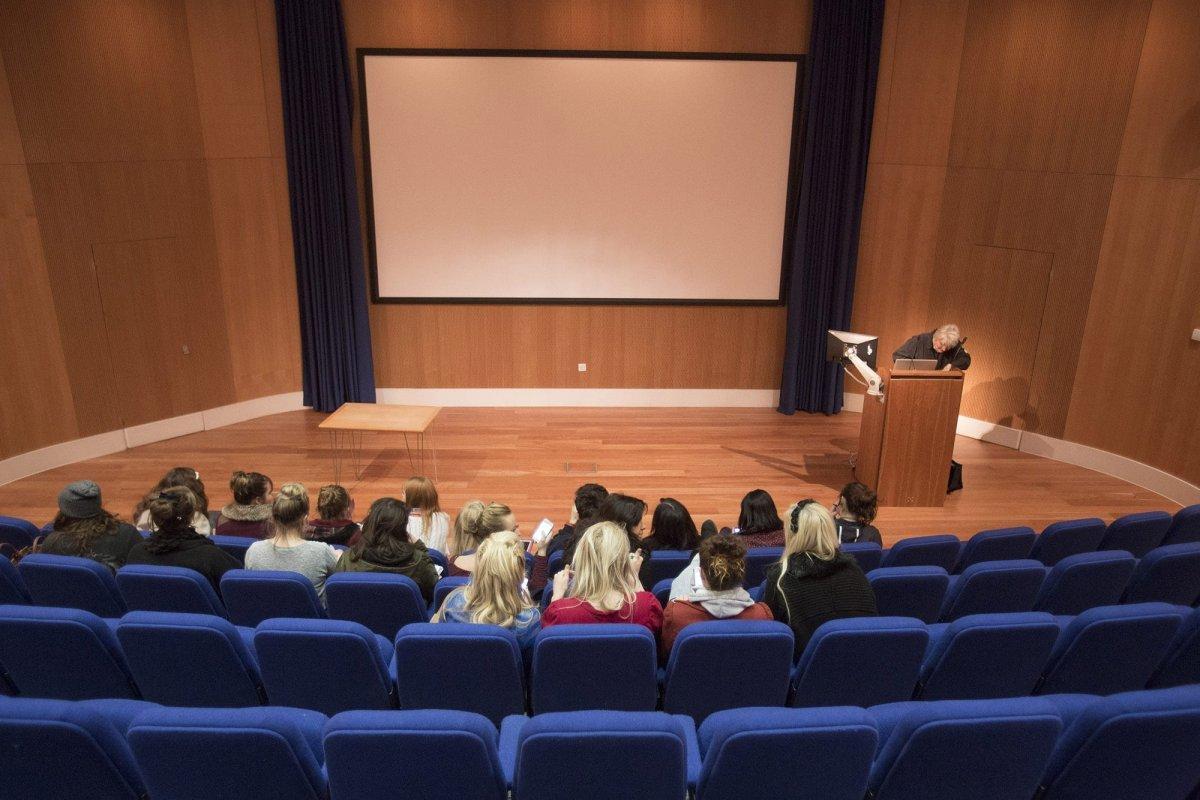 A screen at the front of a lecture theatre. A small group of people is sitting in lower rows of seats.