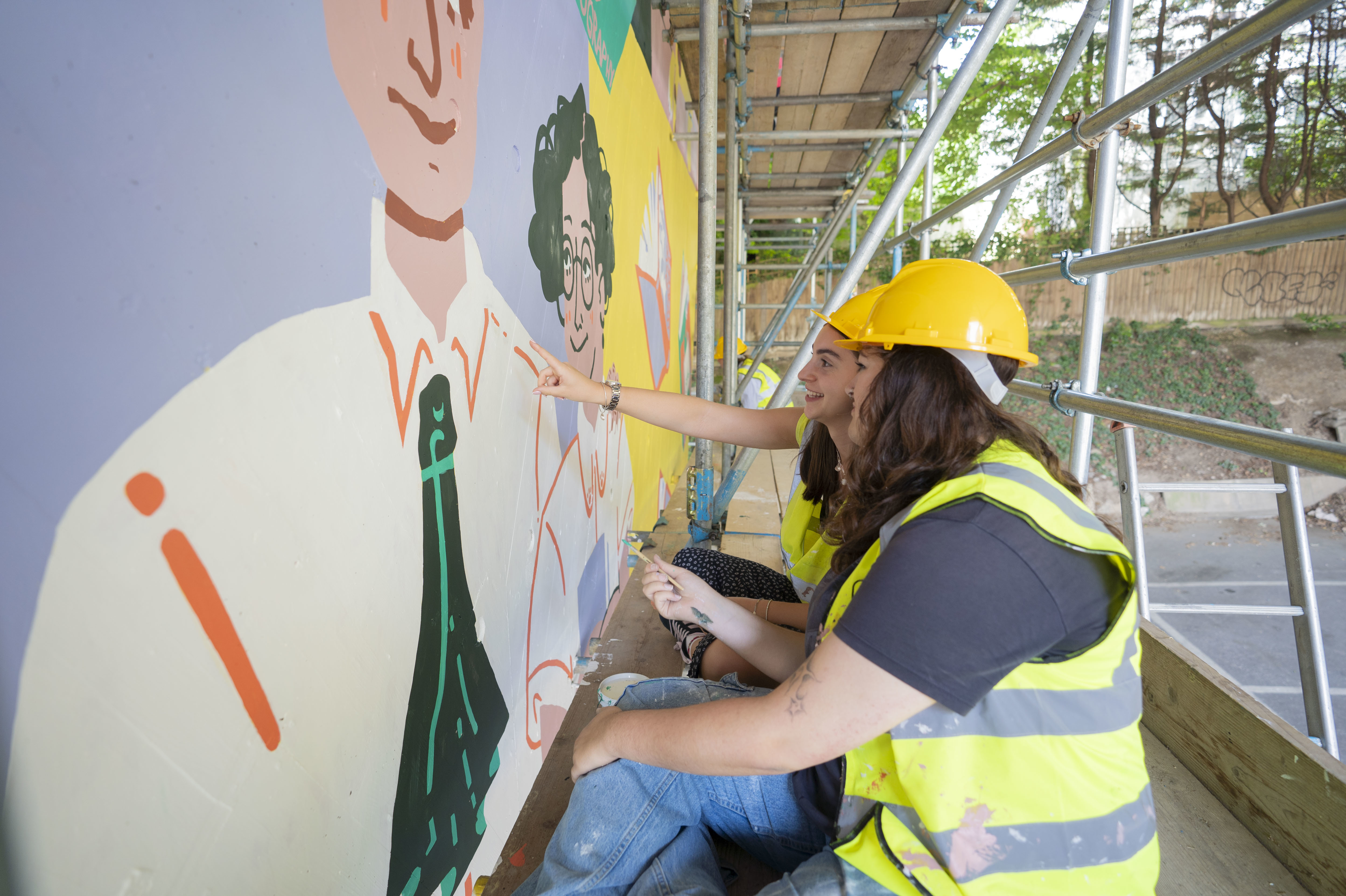 Artists painting the colourful mural at Braidley Road wearing different coloured high-vis, scaffolding.
