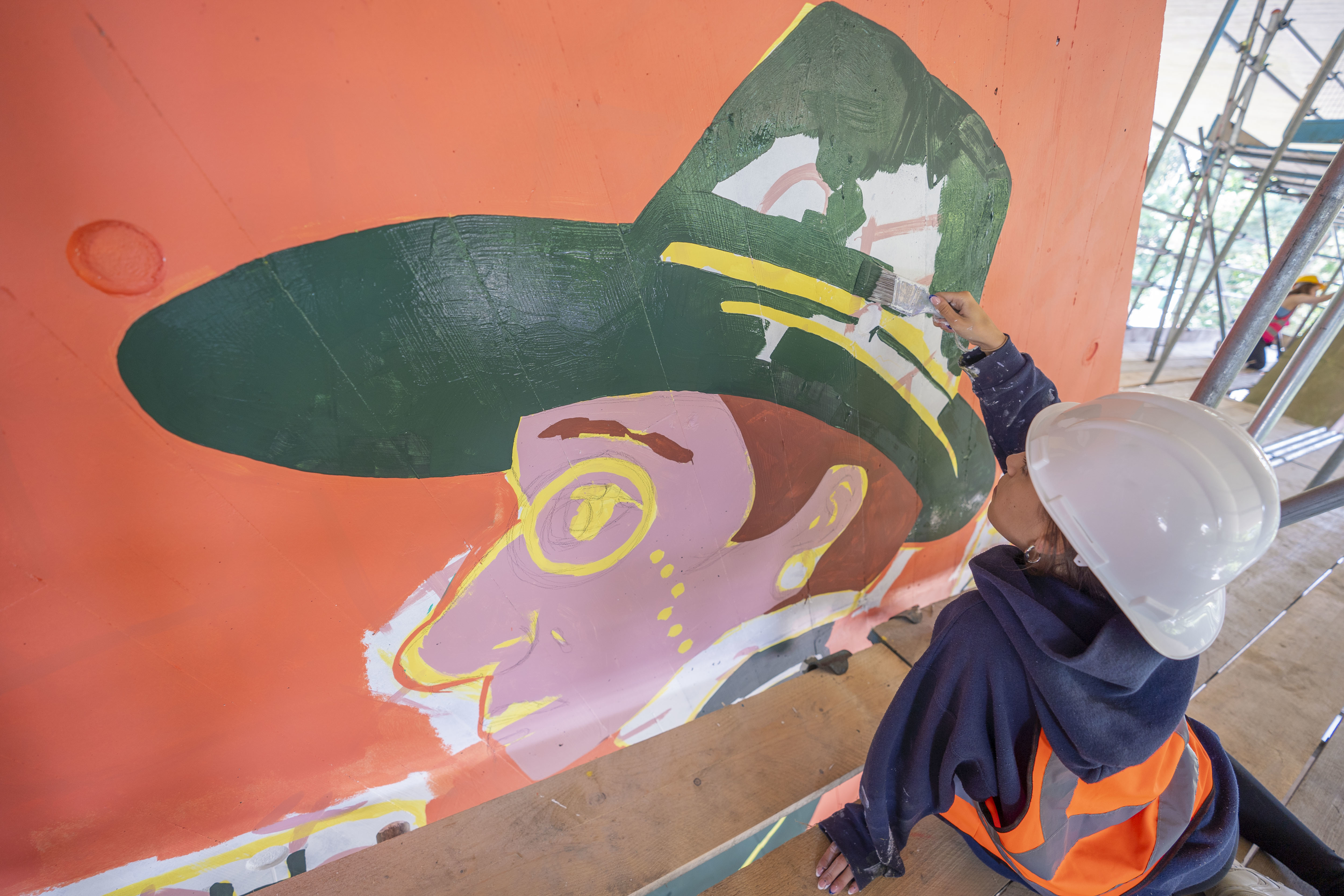 An artist painting a character with a green hat as part of colourful mural at Braidley Road wearing an orange high-vis, scaffolding.