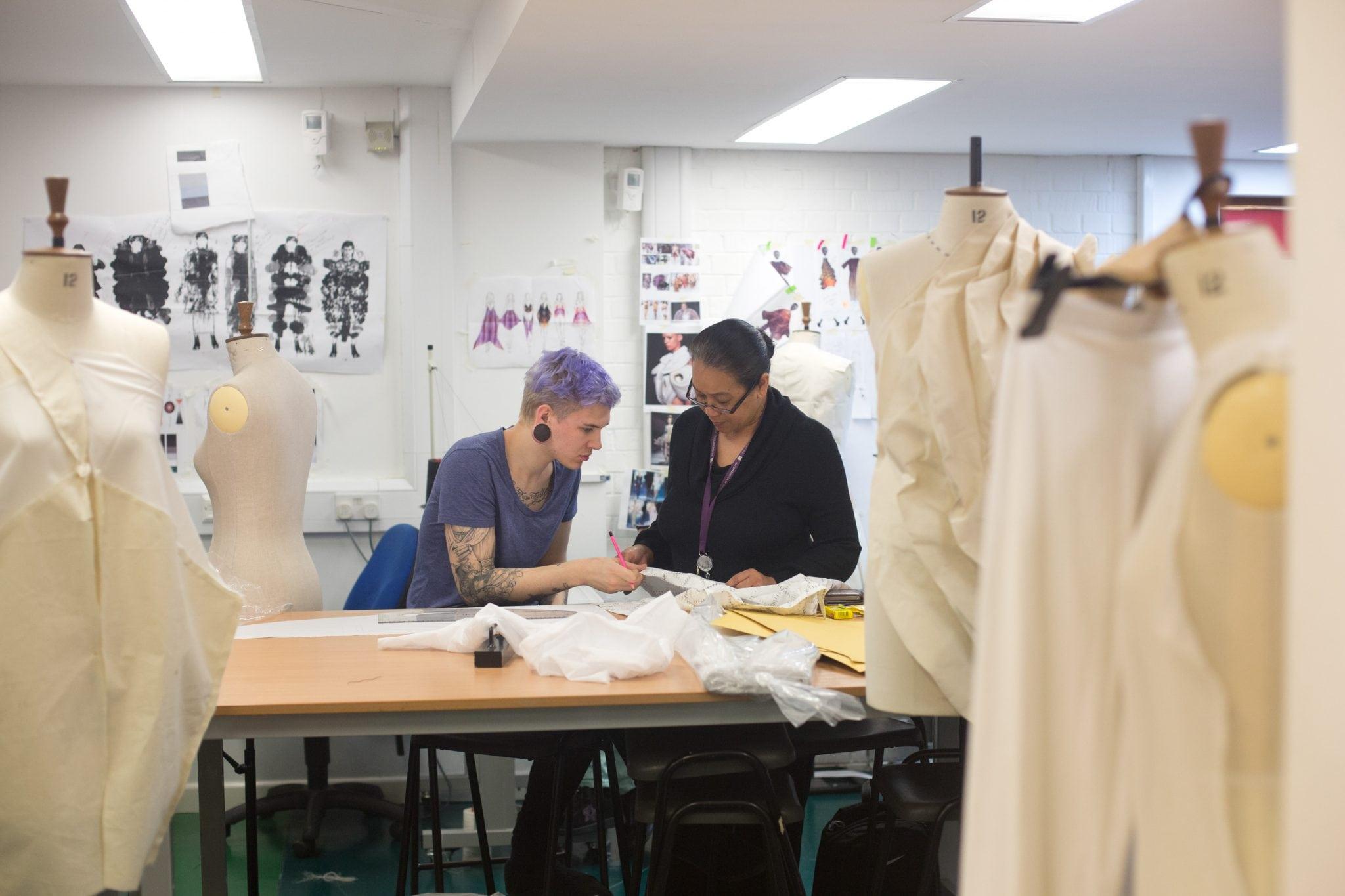 Two students working in a studio sat at a table and surrounded by dressed mannequins.