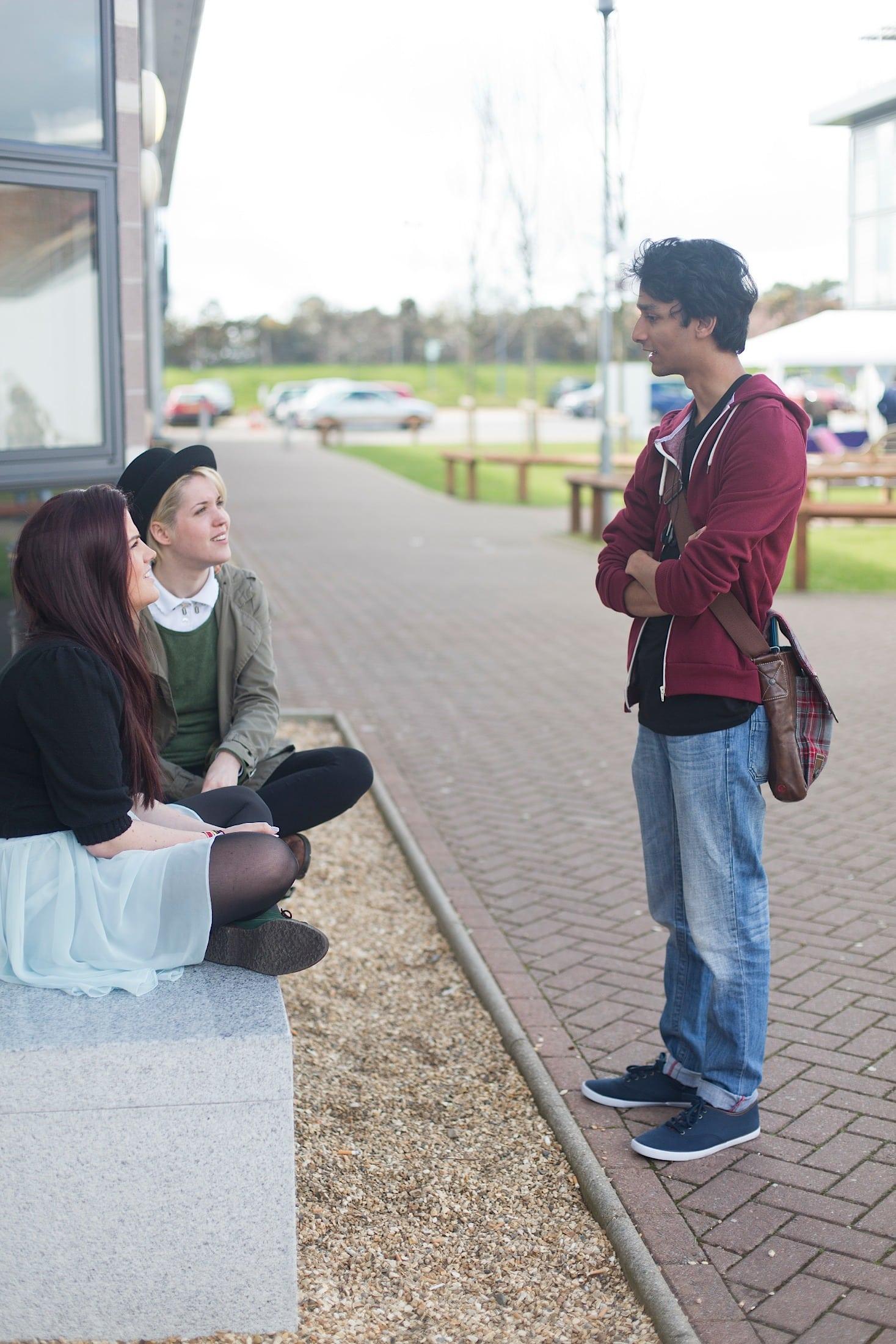 Two students sitting on a bench outside library, chatting with another student who is stood up.