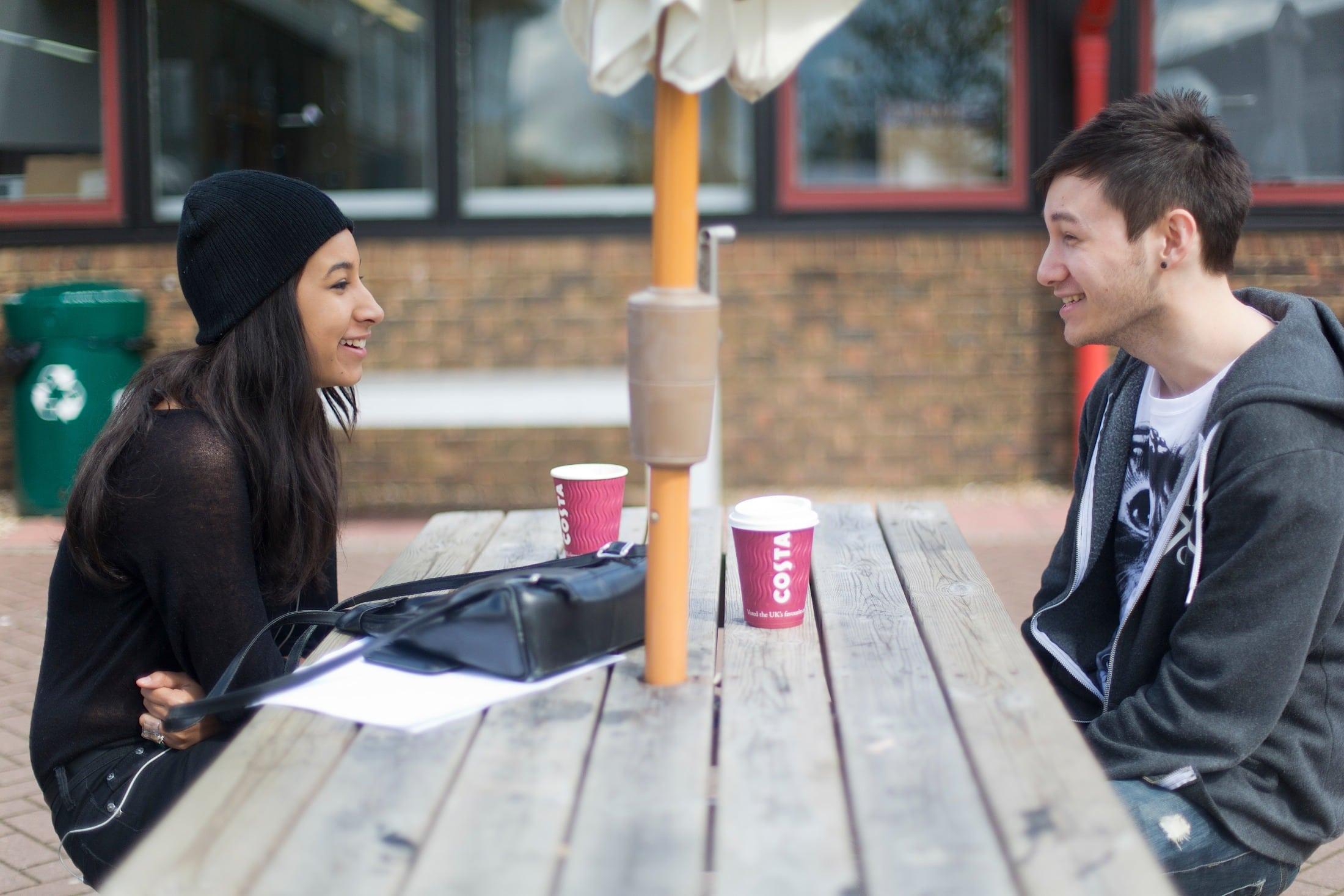 Two students sitting on opposite sides of a picnic bench. Both have coffee cups in front of them.