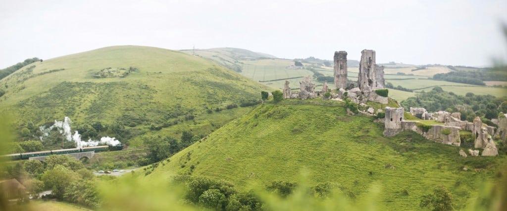A green steam train travelling through green hills and fields. On the right are the old ruins of Corfe Castle