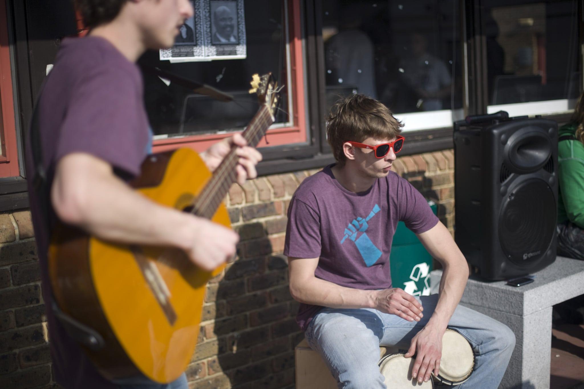 Two musicians performing outside a university building. One is standing playing a guitar. The other is seated and playing bongo drums.