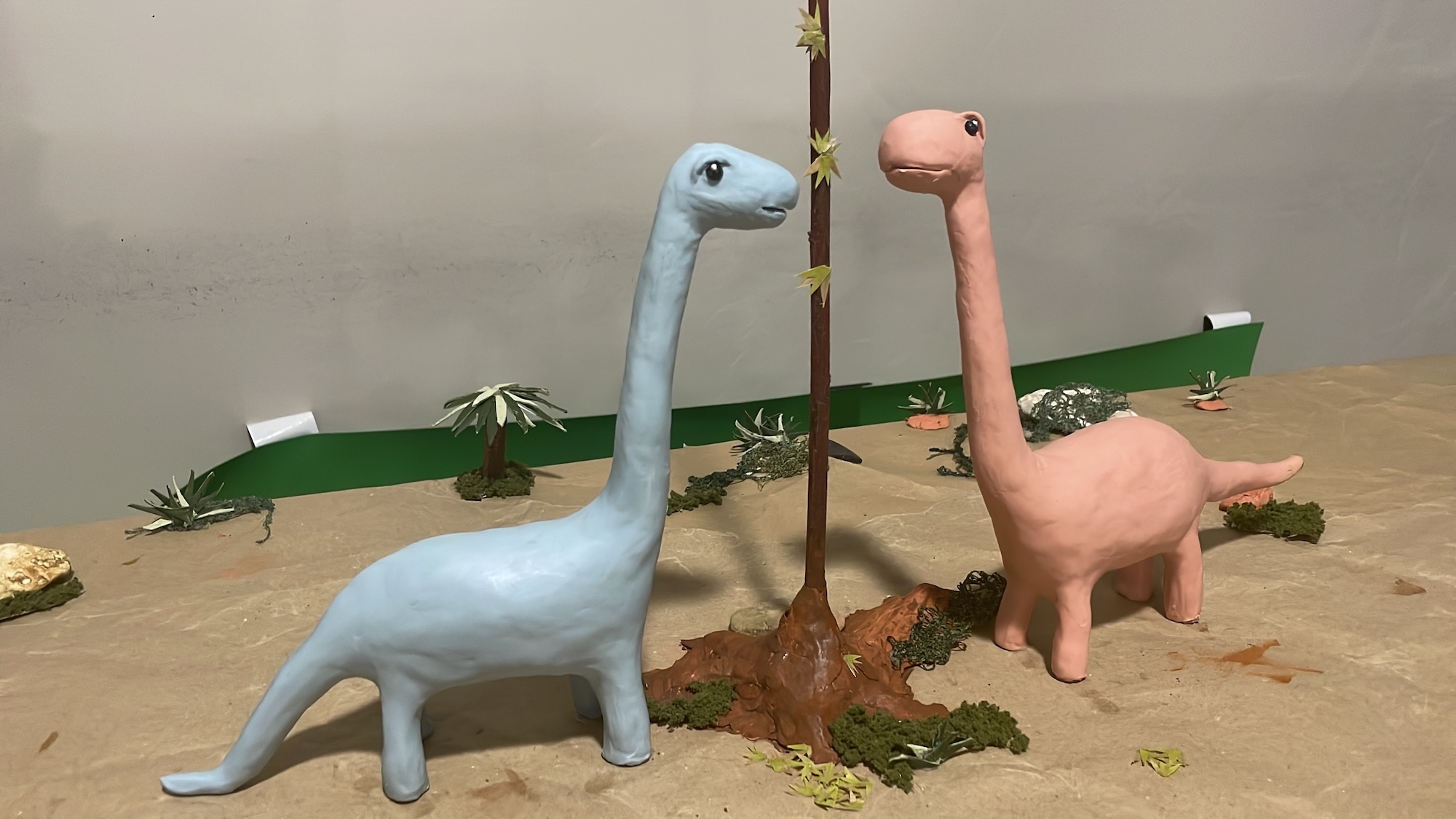 Two animated dinosaur models, one blue, one orange, both looking at a tree with few leaves.