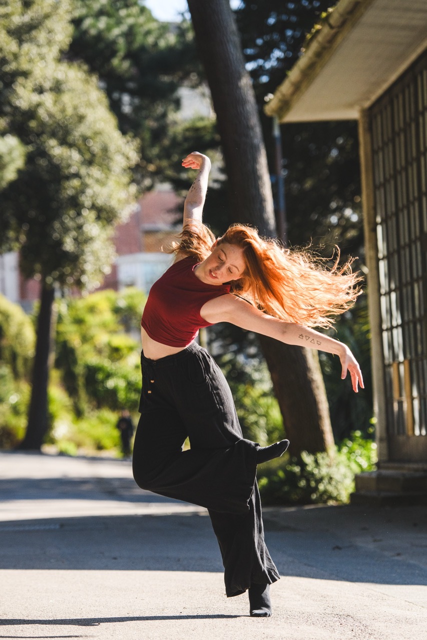 Woman in burgundy top and black trousers mid-dance leap on sunny street, hair flowing, trees and buildings in background.