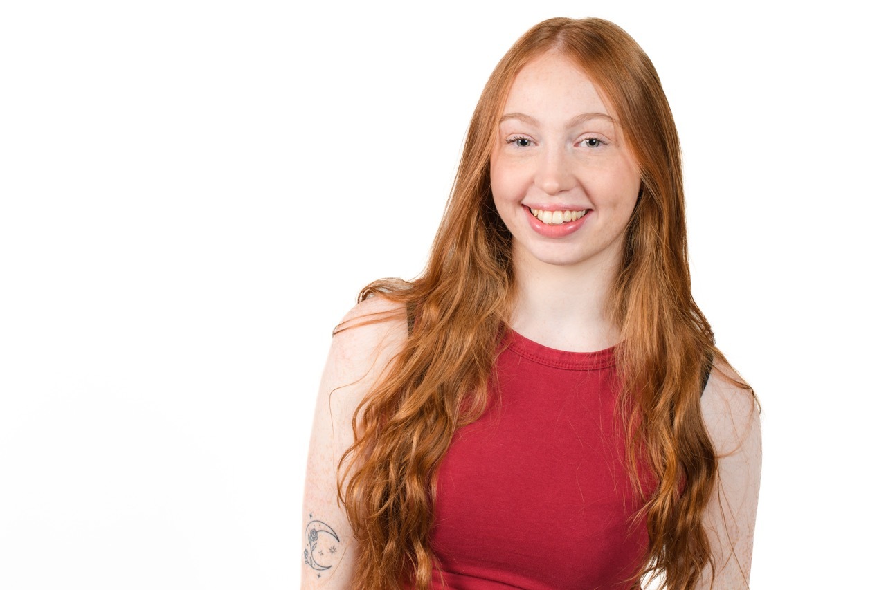 Young woman with long ginger hair wearing red sleeveless top, smiling at camera against white background.