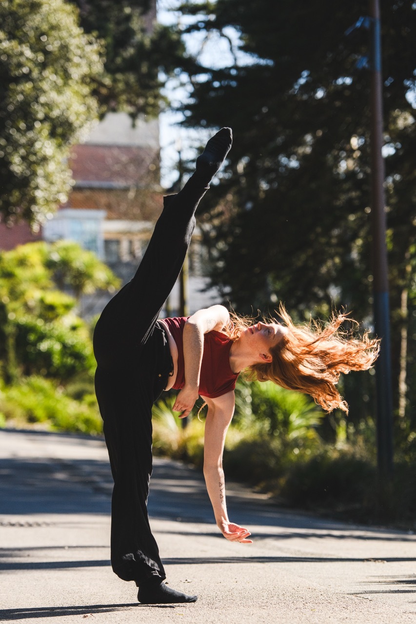 Woman in red top and black leggings performing high kick on pavement, surrounded by green trees and buildings in background.