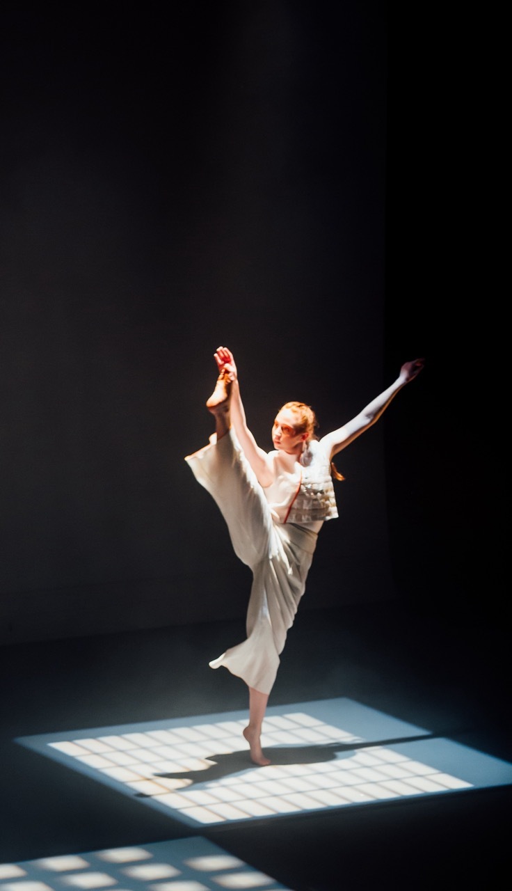Ballet dancer in white costume performing high leg extension on illuminated square stage against black background.