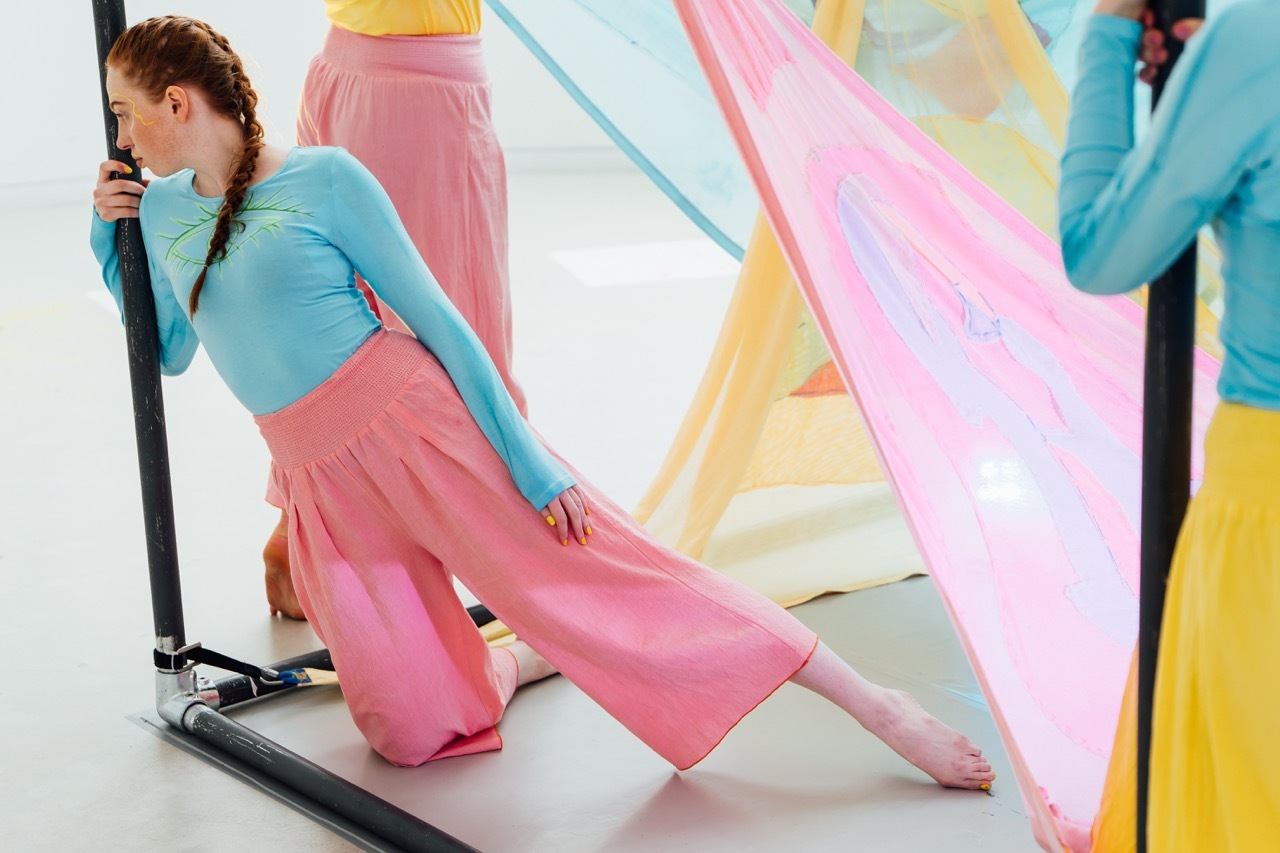 Woman in blue top and pink trousers kneeling on reformer equipment amongst colourful translucent fabric drapes in pastels.