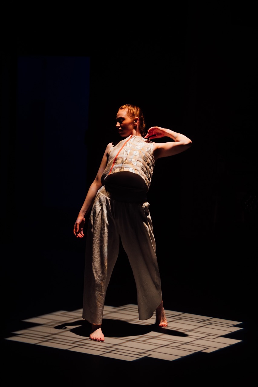 Dancer on chequered mat against dark blue backdrop, wearing white patterned top and grey trousers, one hand raised to head.