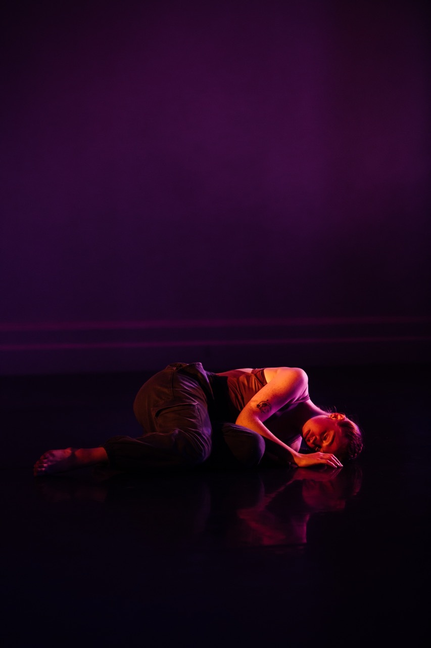 Person lying on reflective black surface illuminated by purple, pink and orange lighting against dark purple background.