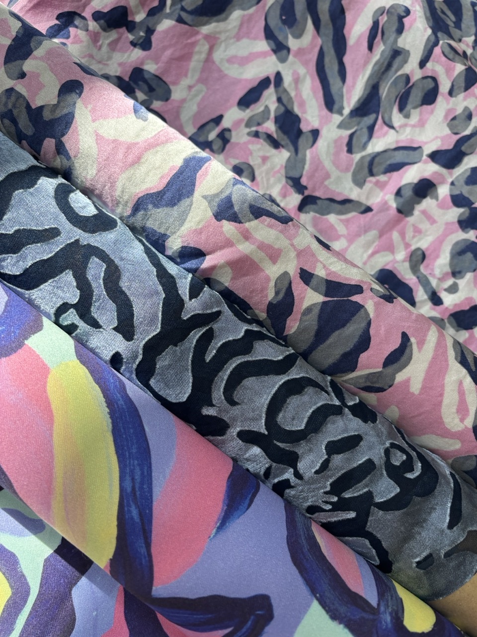Colourful fabric with camouflage patterns in pink, grey, and navy blue arranged in flowing, overlapping folds and curves.