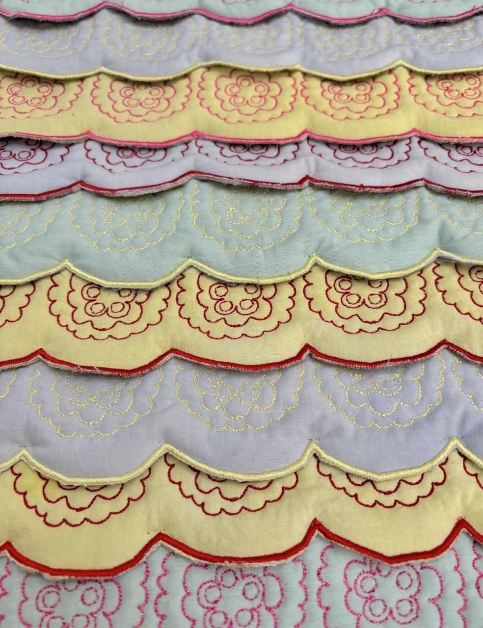 Horizontal rows of scalloped fabric trim in yellow, lilac, and white with red floral embroidery and decorative stitching.