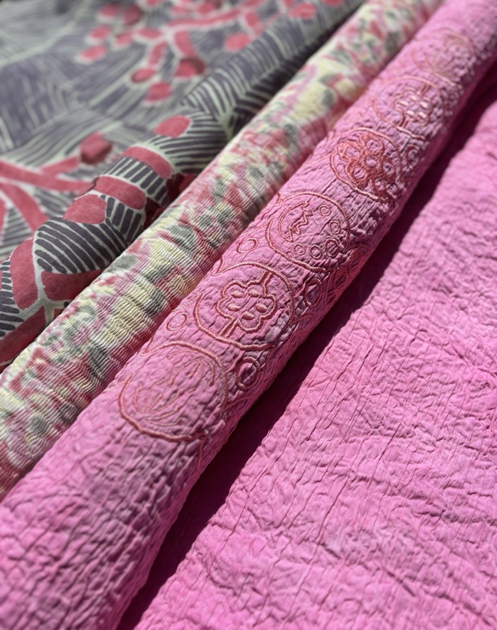 Folded textiles in pink and patterned fabrics with floral prints, geometric designs, and embossed textures arranged diagonally.