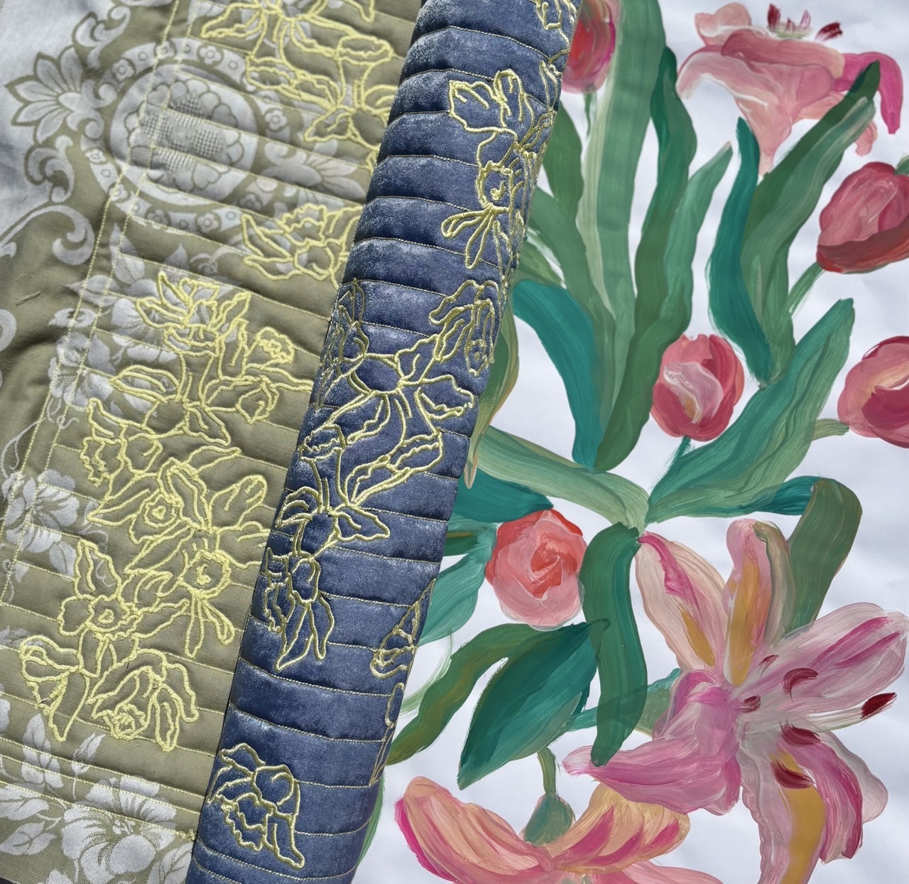 Quilted fabric with yellow embroidered florals on grey and blue sections alongside painted pink and coral flowers with green stems.