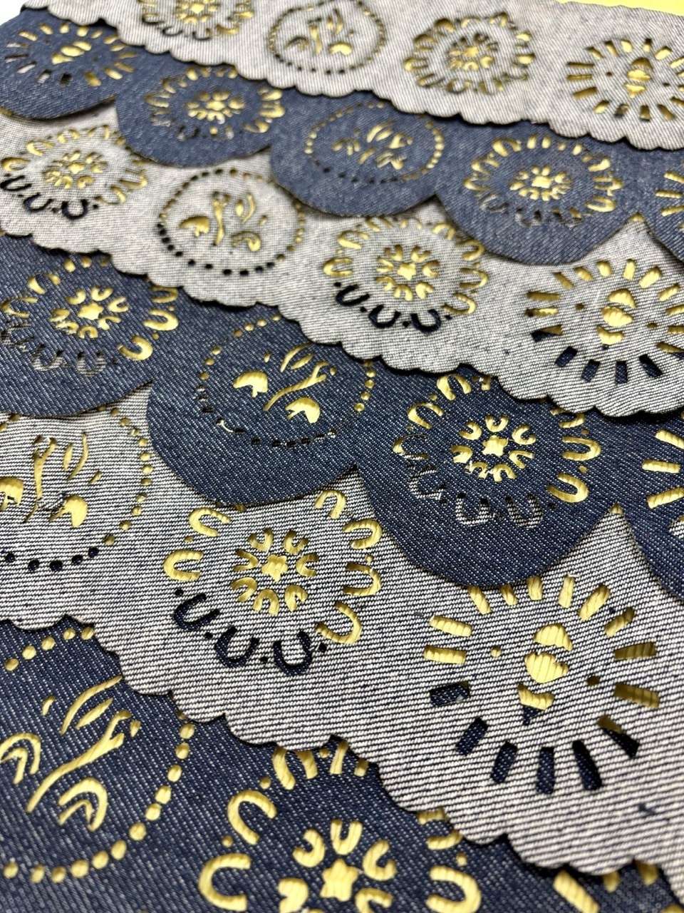 Embroidered fabric with circular floral motifs in yellow and black on blue and grey backgrounds, featuring scalloped edges and layered composition.