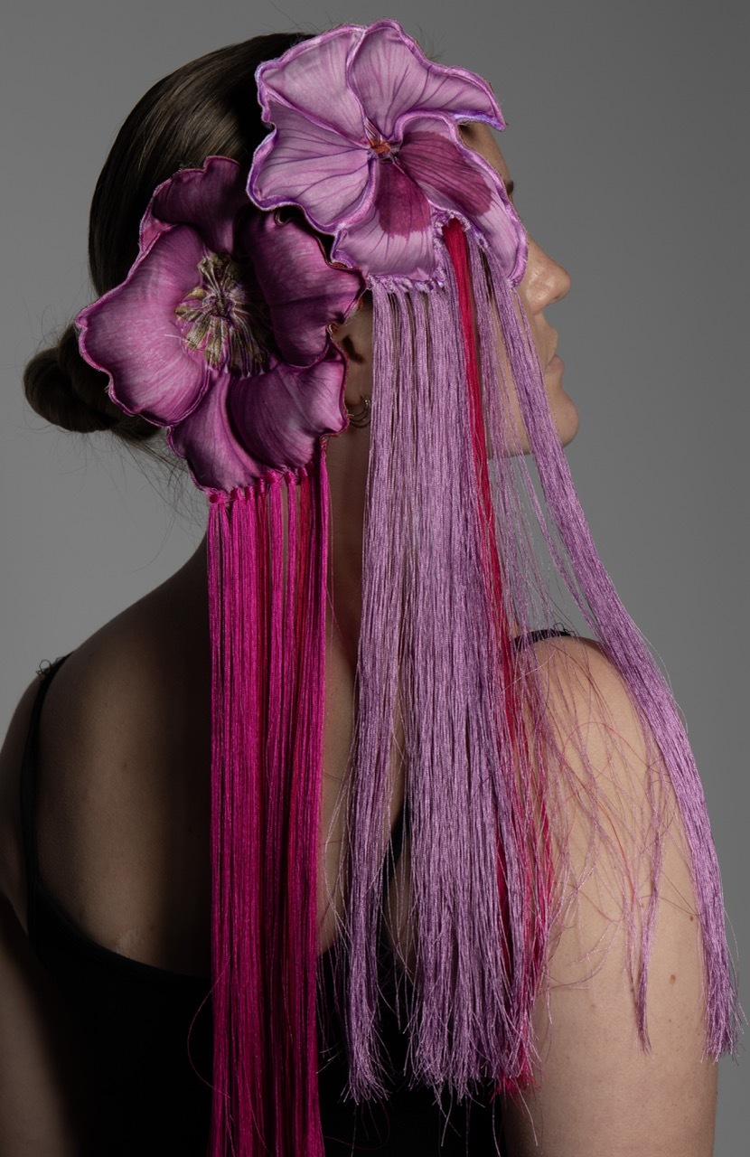 Woman in profile wearing large purple fabric flowers in her dark hair with cascading purple and magenta tassels against grey background.