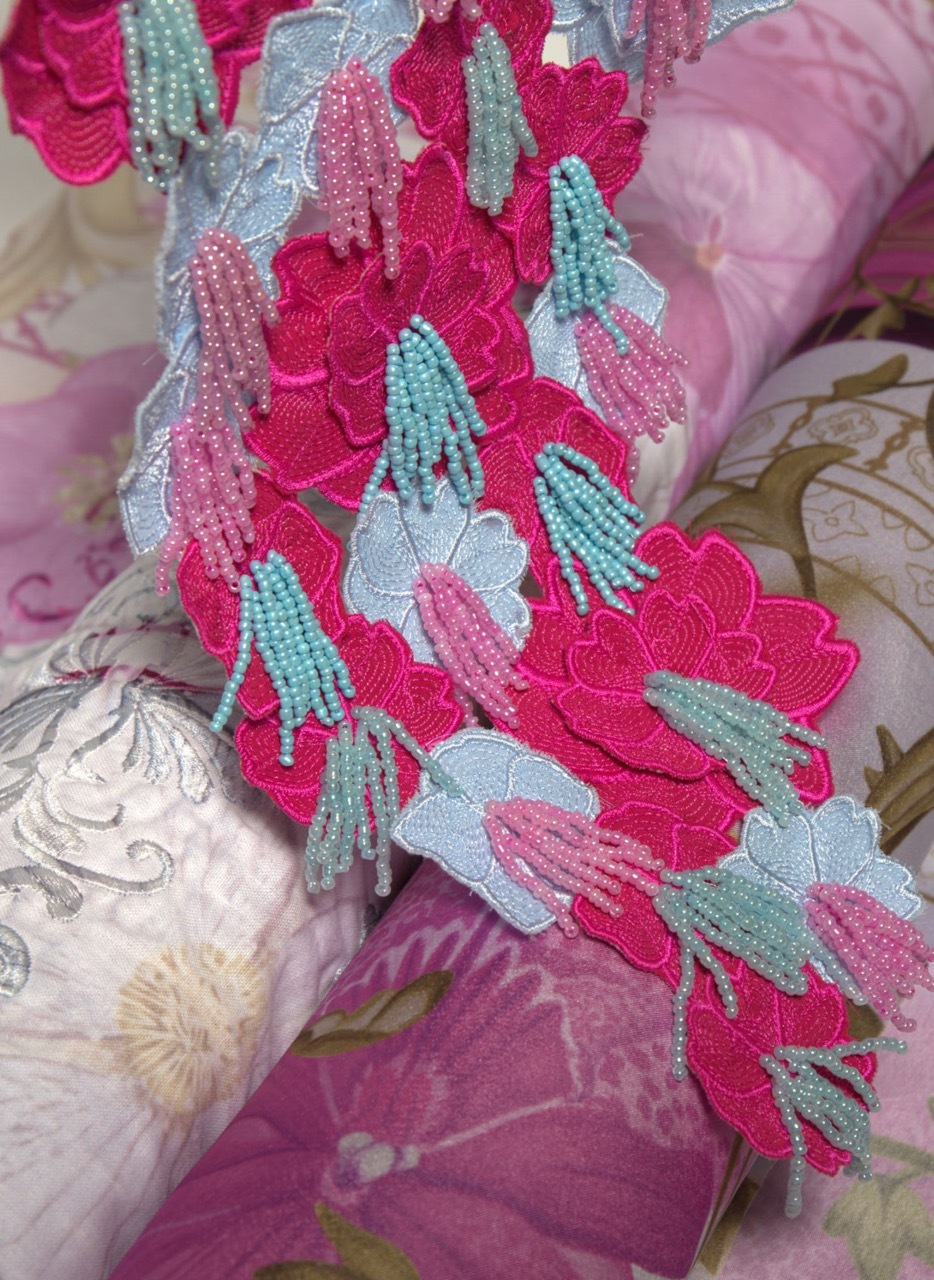 Intricate beadwork with bright pink flowers, pale blue leaves, and mint green tassels on floral fabric background.