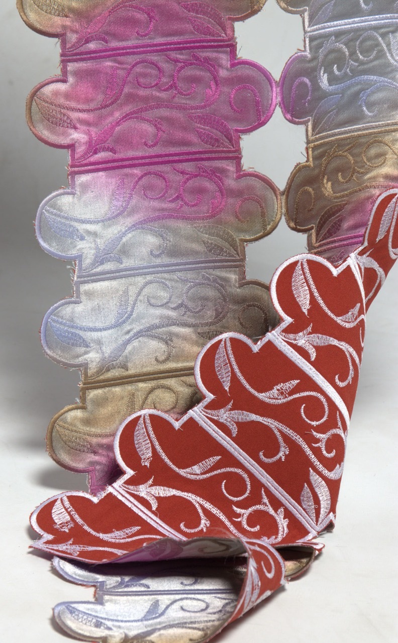 Stacked ornate curved shapes in pink, blue, and brown tones with decorative swirls, topped by red ribbon-like forms with white scroll patterns.