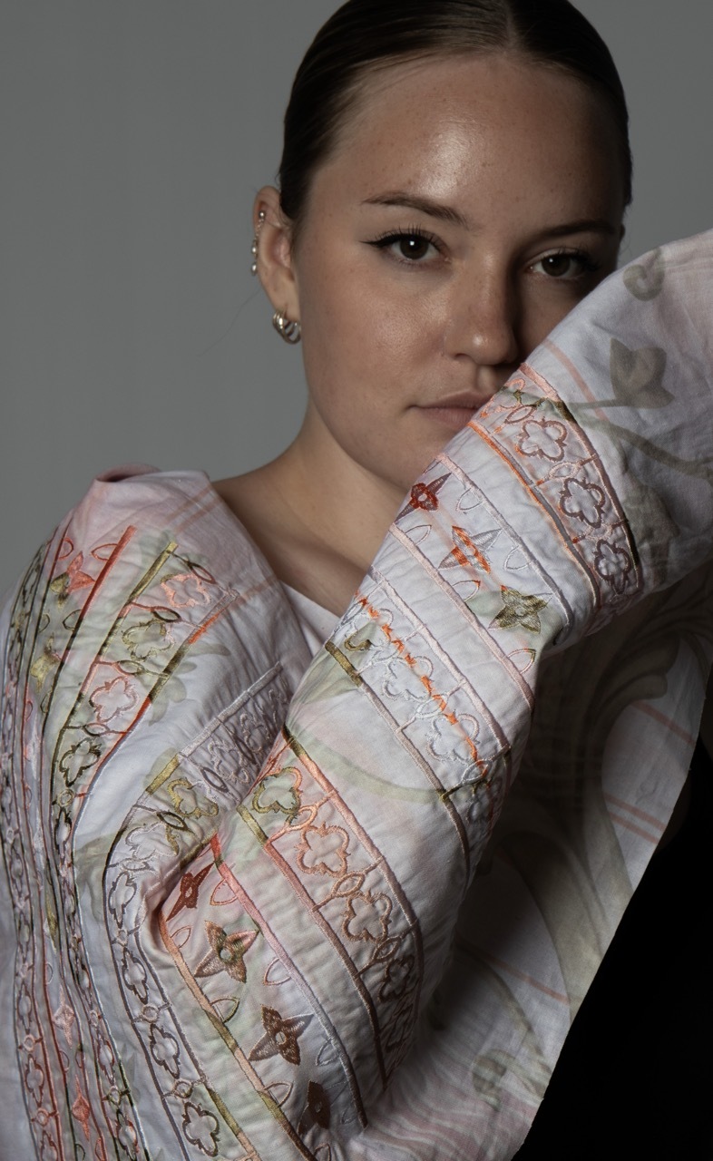 Woman with slicked-back hair wearing white silk shirt with colourful floral and geometric patterns, hand positioned near face.