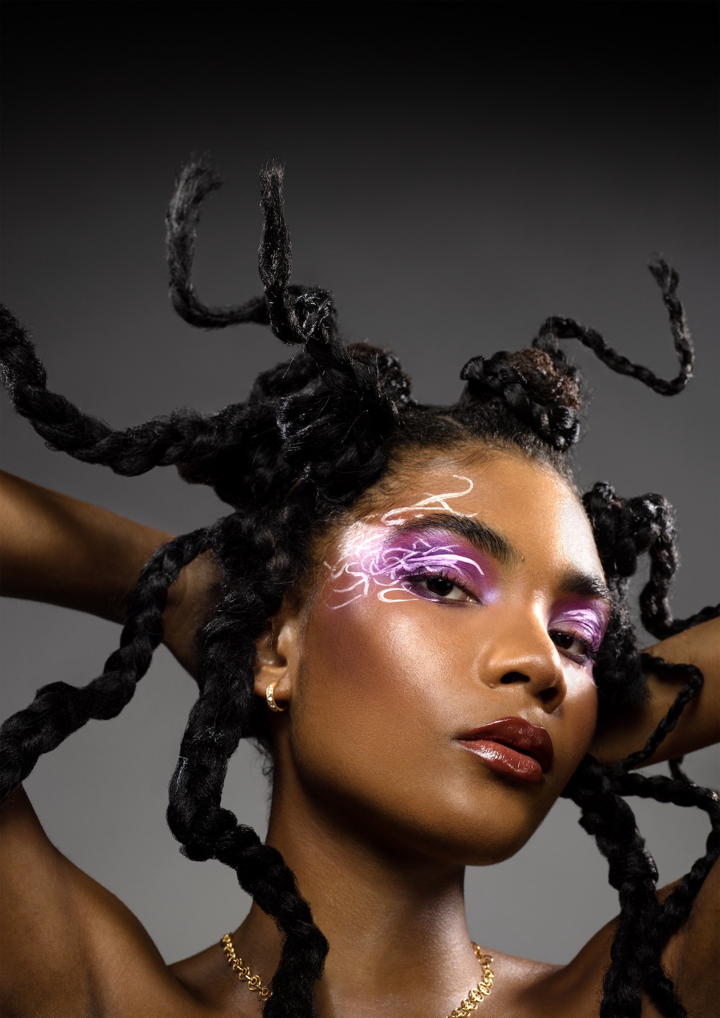 Headshot of a model with flashy eyeshadow, looking towards the camera, posing with their hands behind their head.