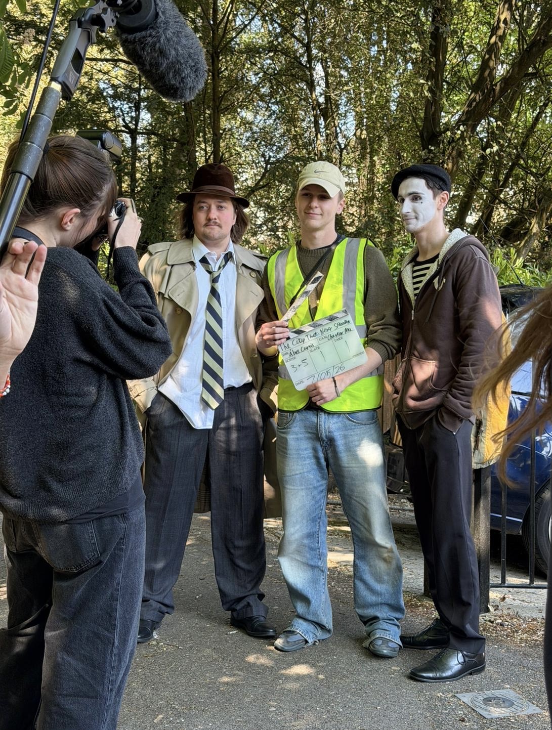 A film crew stands outdoors, with a man in a trench coat and hat, a man in a yellow safety vest holding a clapperboard, and a man with white face paint. Surrounding them are crew members and trees in the background.
