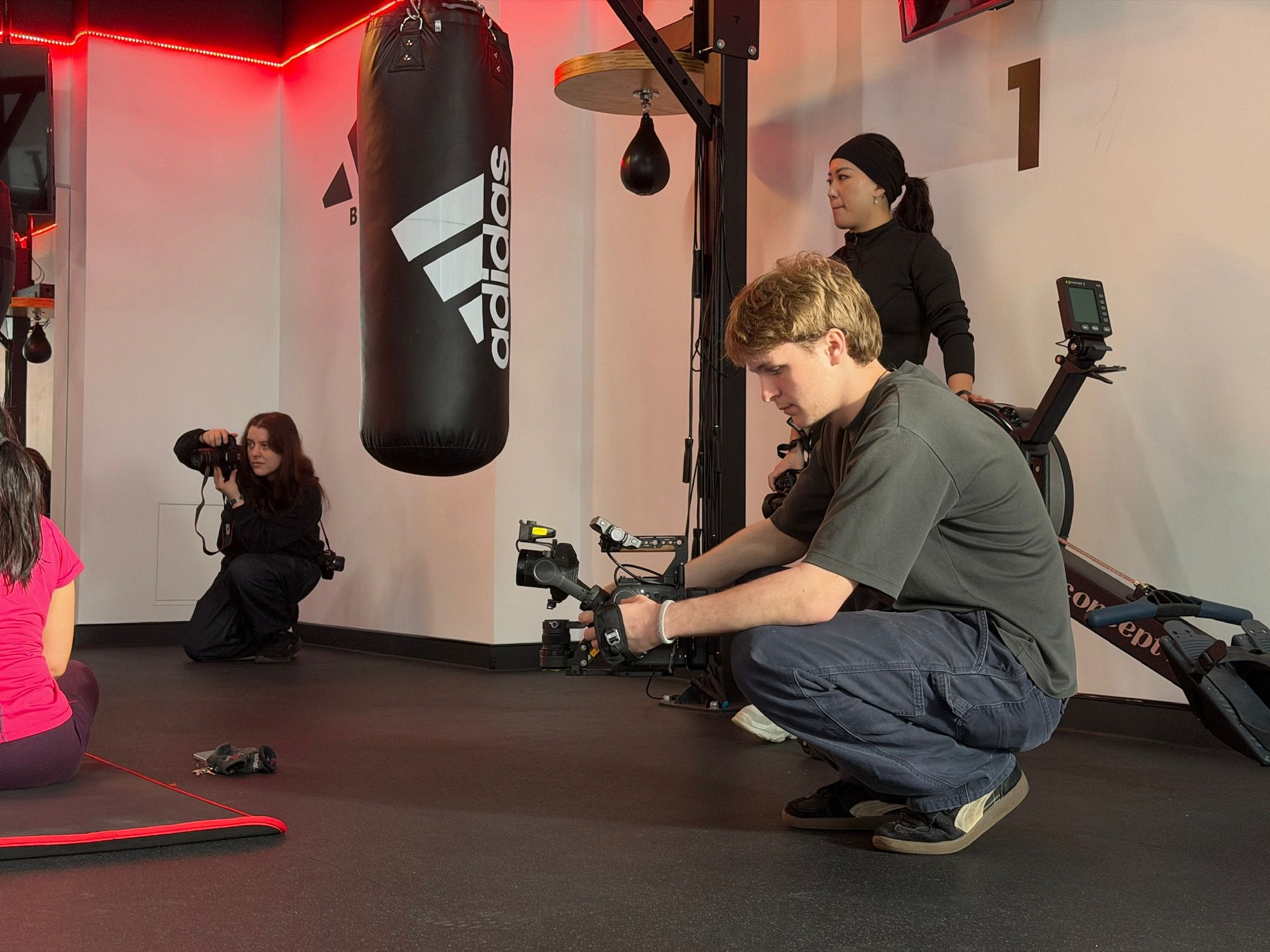 BA (Hons) Film Production student A. Curtis crouches while filming with a camera in a gym setting. In the background, a woman with long hair stands next to a punching bag, and another woman is seated on a mat. The space features red lighting and exercise equipment.