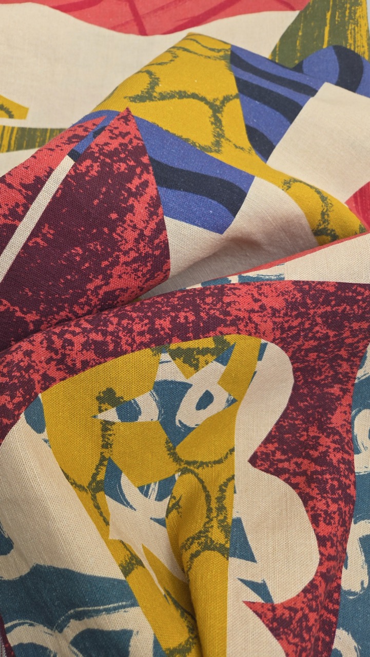 A folded piece of fabric featuring an abstract design with bold shapes in vibrant colours, including red, yellow, blue, and green. The texture of the material is visible, showcasing a mix of patterns.